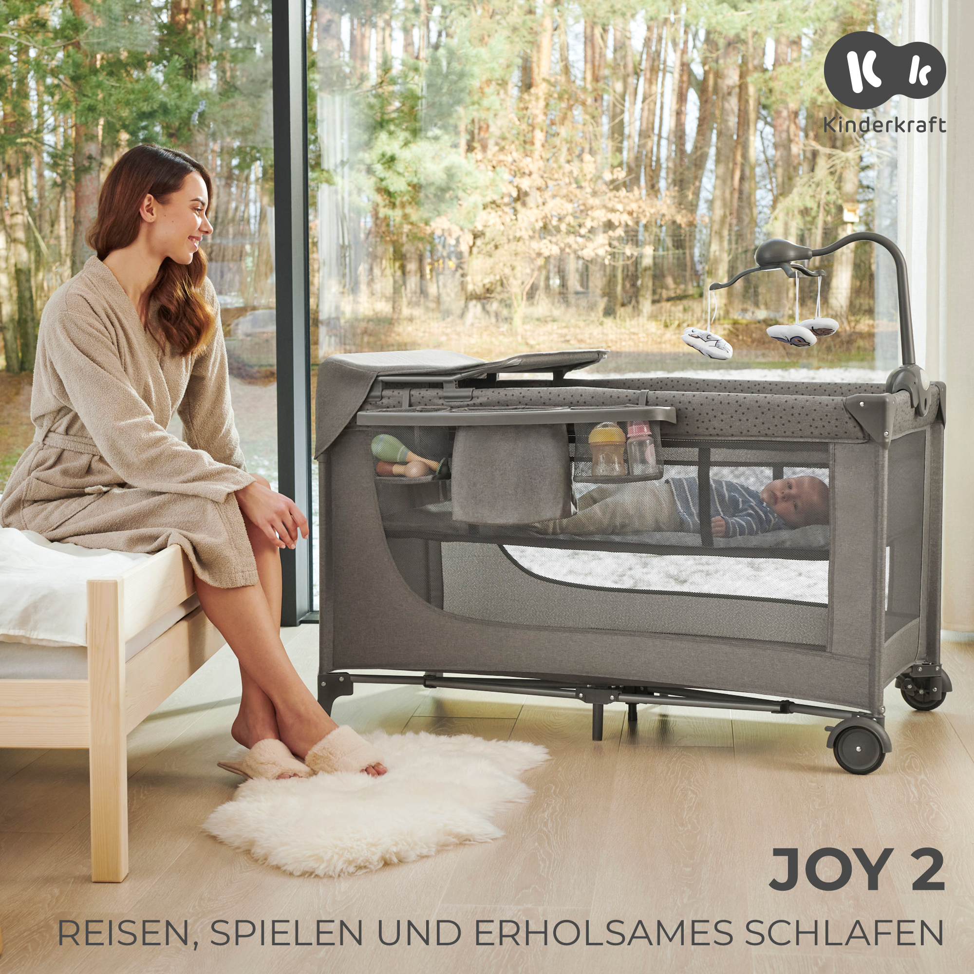 Eine Frau in einem Bademantel sitzt auf einem Bett neben einem Reisebett mit einem Baby darin.