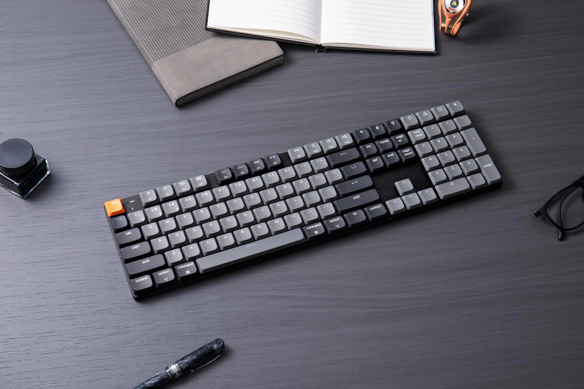 Czarna klawiatura z szarymi klawiszami, pomarańczowym klawiszem ESC, na ciemnym drewnianym biurku.