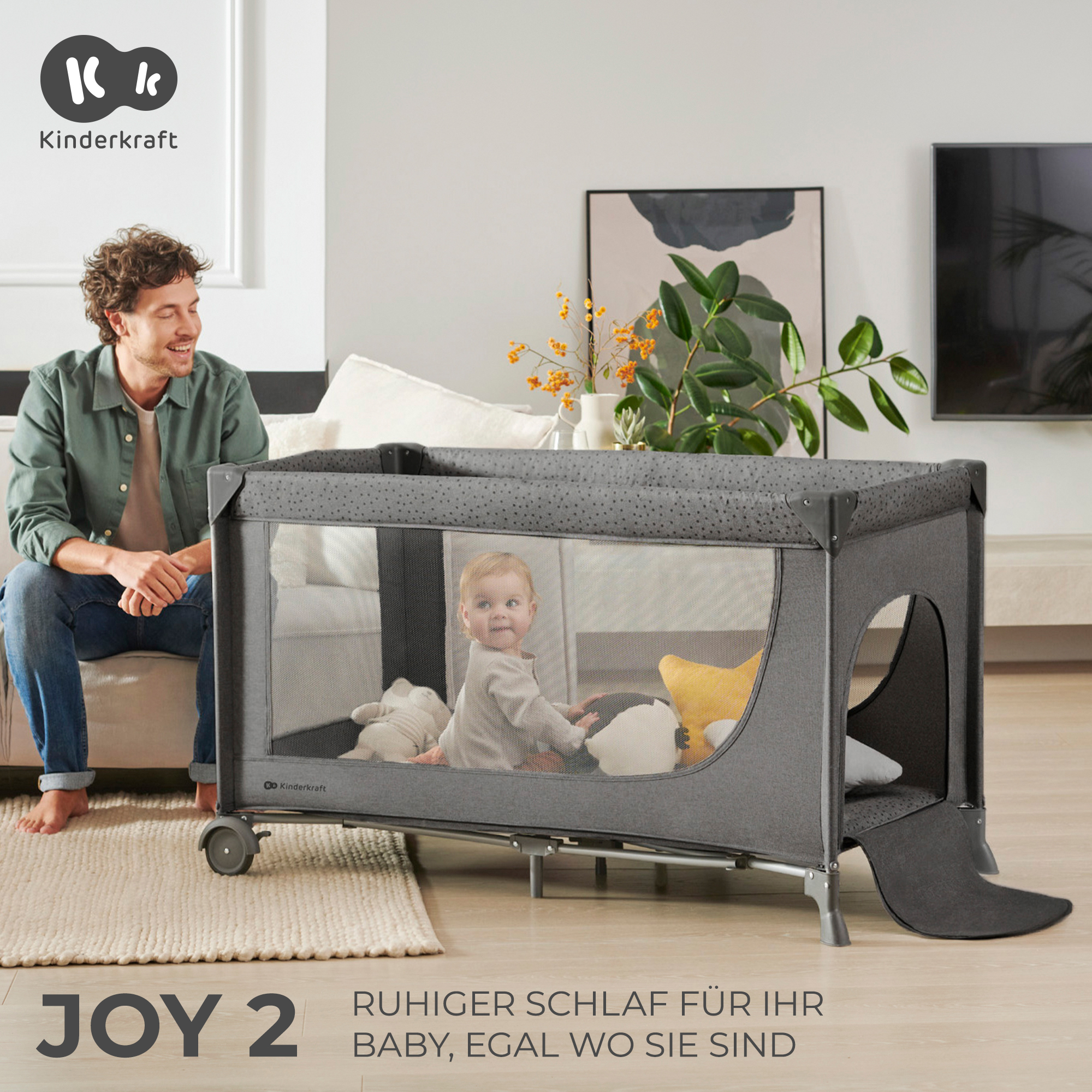 Ein Mann sitzt neben einem grauen Kinderreisebett, in dem ein Baby spielt.