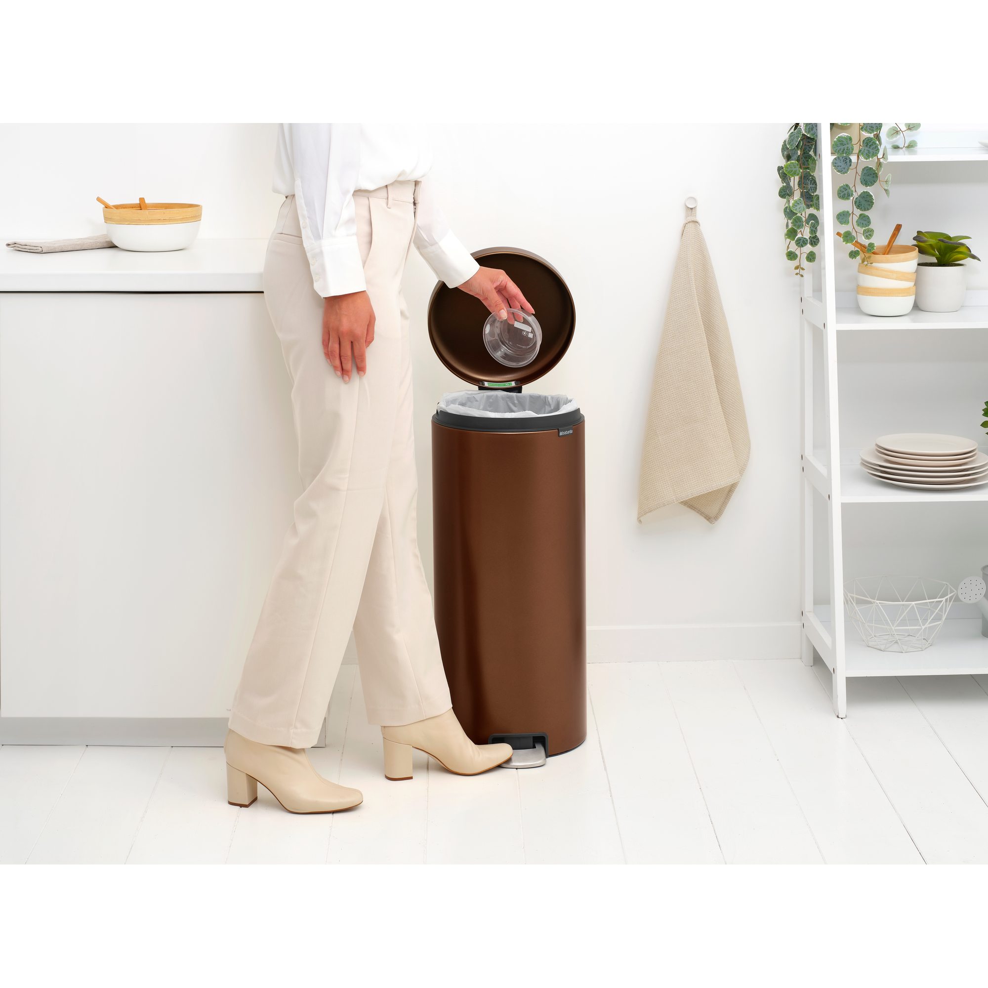 A woman discards a glass cup into a bronze step bin in a kitchen.
