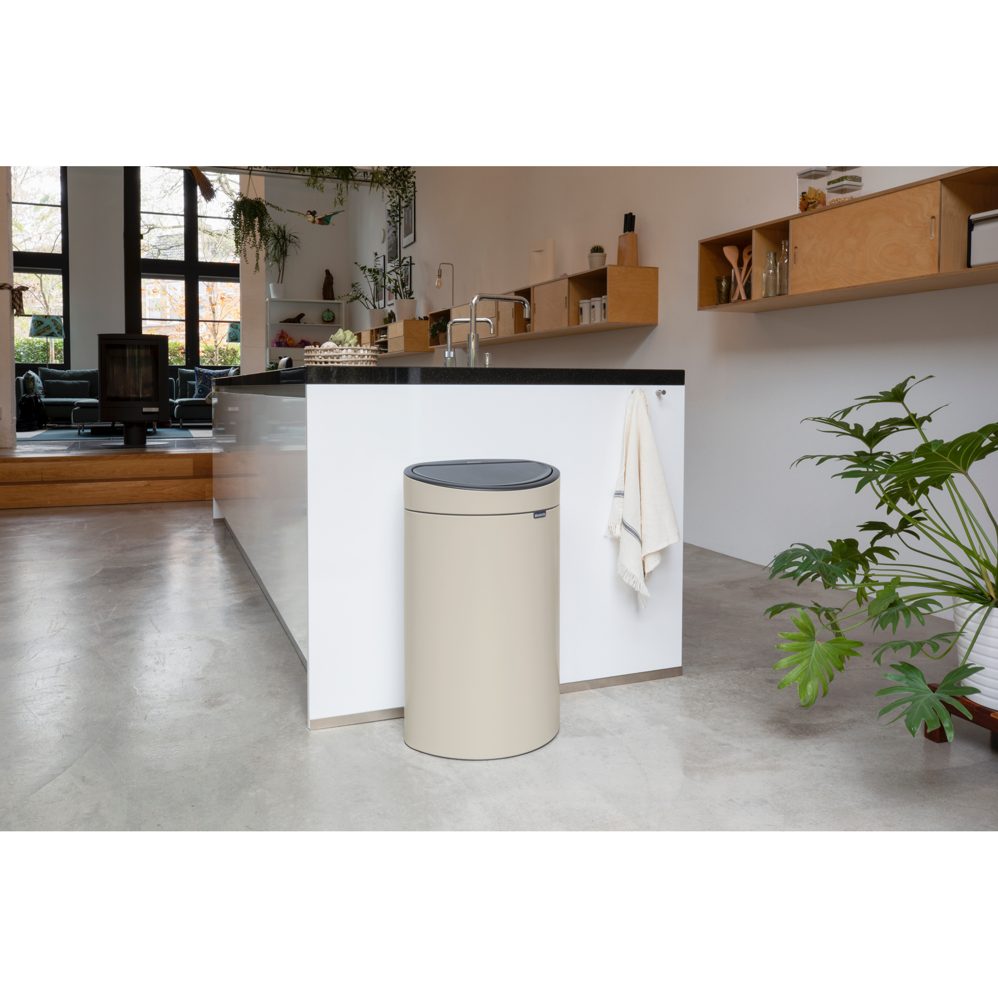 A beige trash can with a black lid sits on a concrete floor next to a white wall.