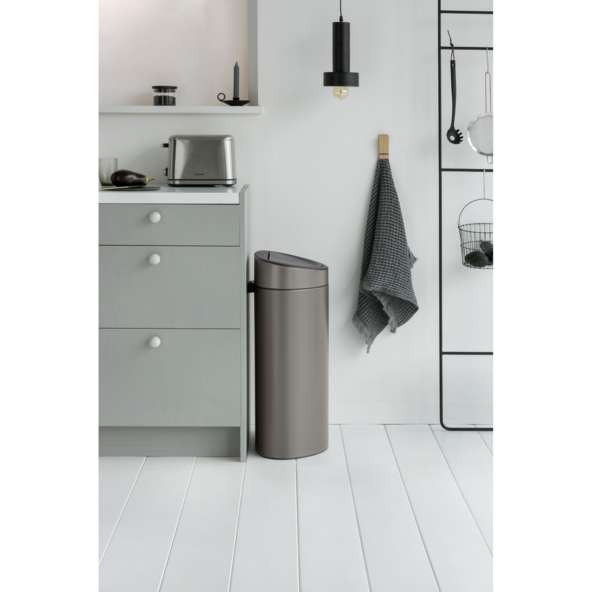 Gray trash can next to a kitchen cabinet with a toaster and eggplant.