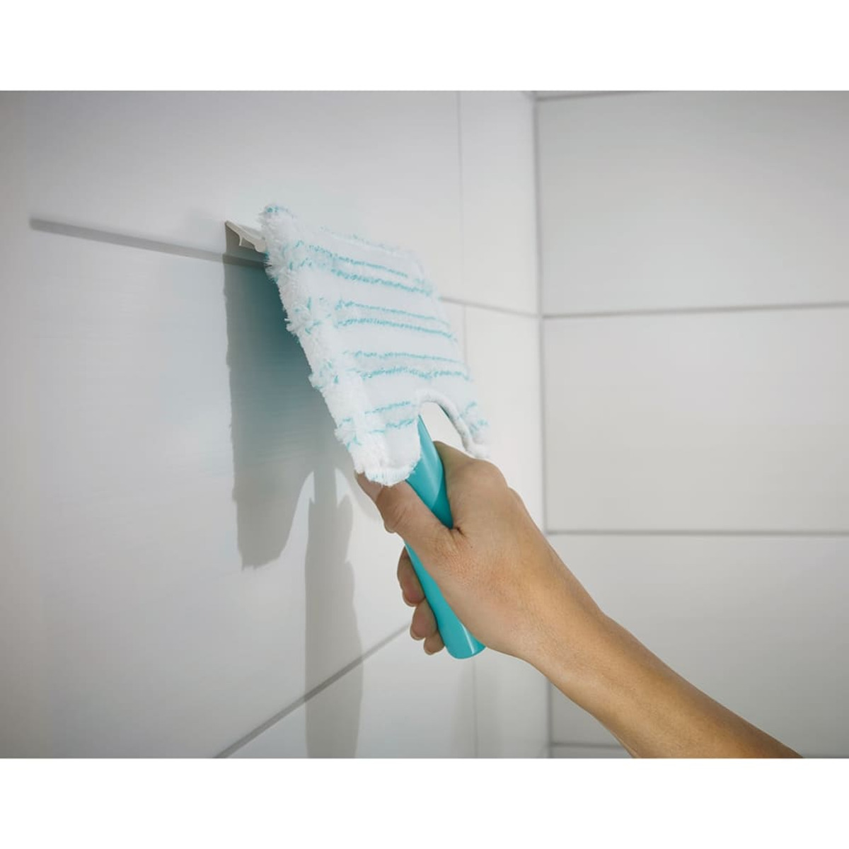 Eine Hand hält einen blauen Griff mit einem weißen und hellblauen Reinigungswerkzeug an einer weißen gefliesten Wand.