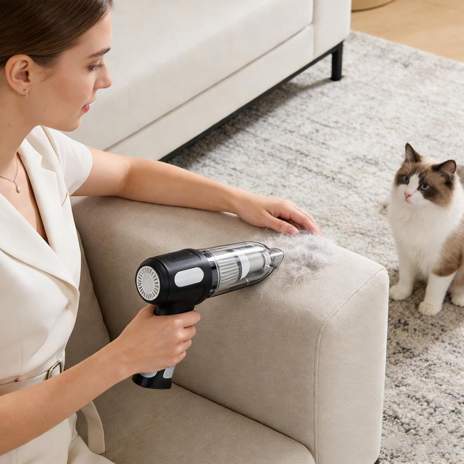 Frau saugt Katzenhaare von einem beigen Sofa. Eine Katze schaut in der Nähe auf einem Teppich zu.