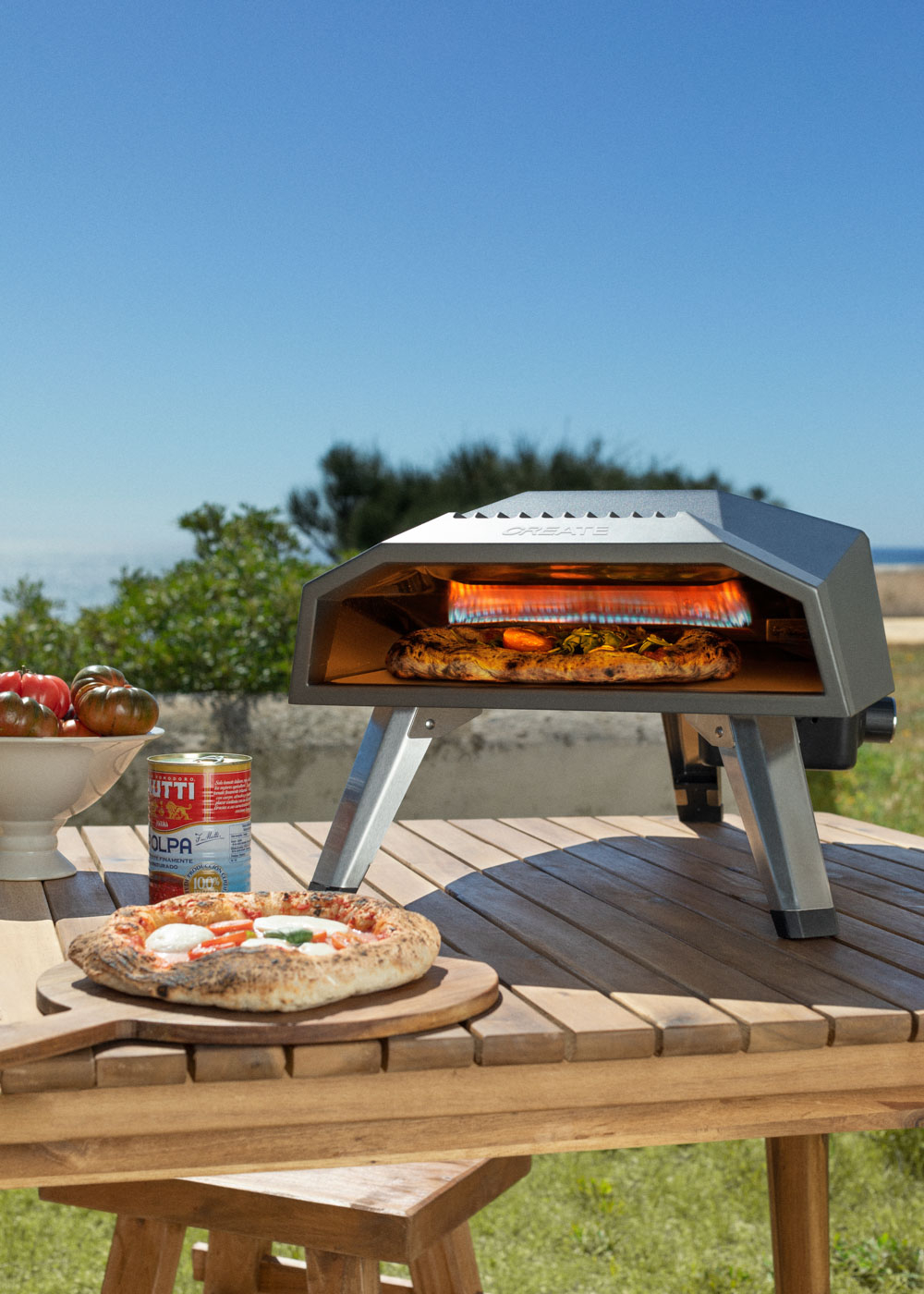 Una pizza se cocina en un horno portátil sobre una mesa de madera. Una pizza terminada está cerca.