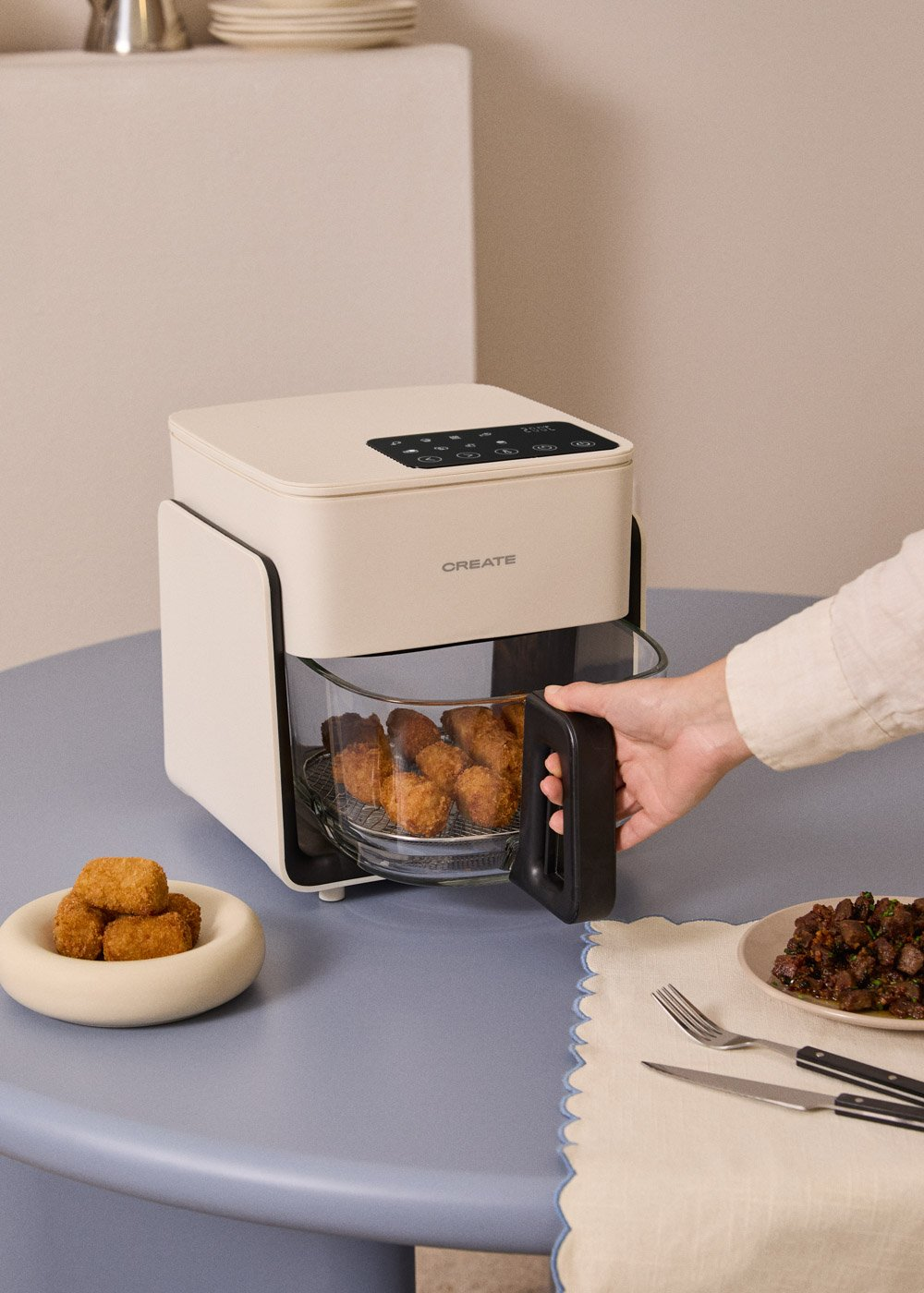 Una freidora de aire blanca con nuggets dorados en la cesta. Hay nuggets y cubiertos en la mesa.