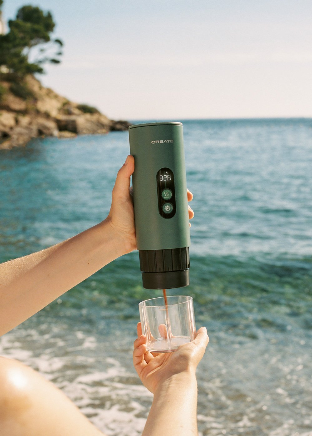 Una cafetera portátil verde prepara café en un vaso en la playa.