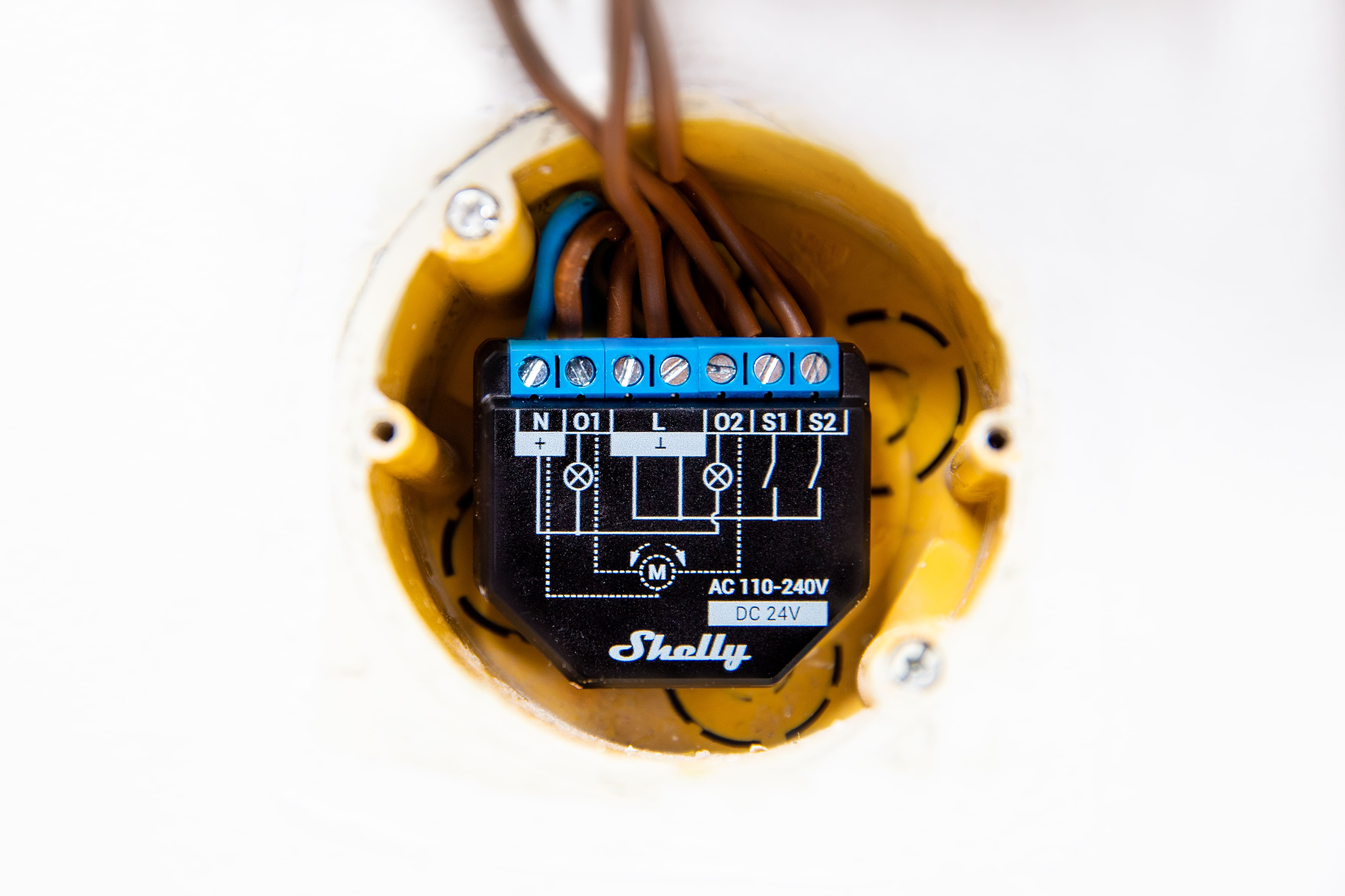 Inteligentne przekaźniki Shelly z podłączonymi przewodami w żółtej puszce elektrycznej.