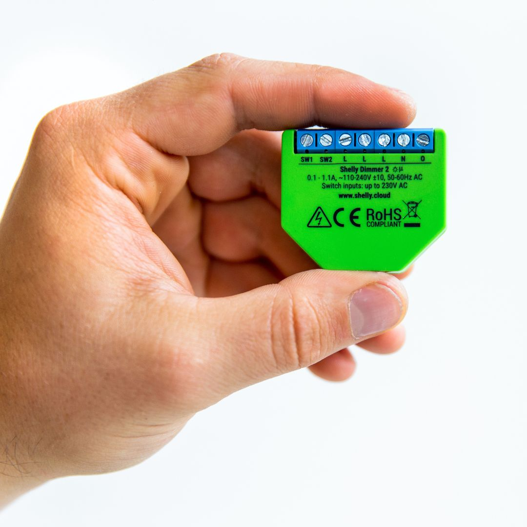 Una mano tiene un dispositivo verde Shelly Dimmer 2 con morsetti blu e testo bianco.