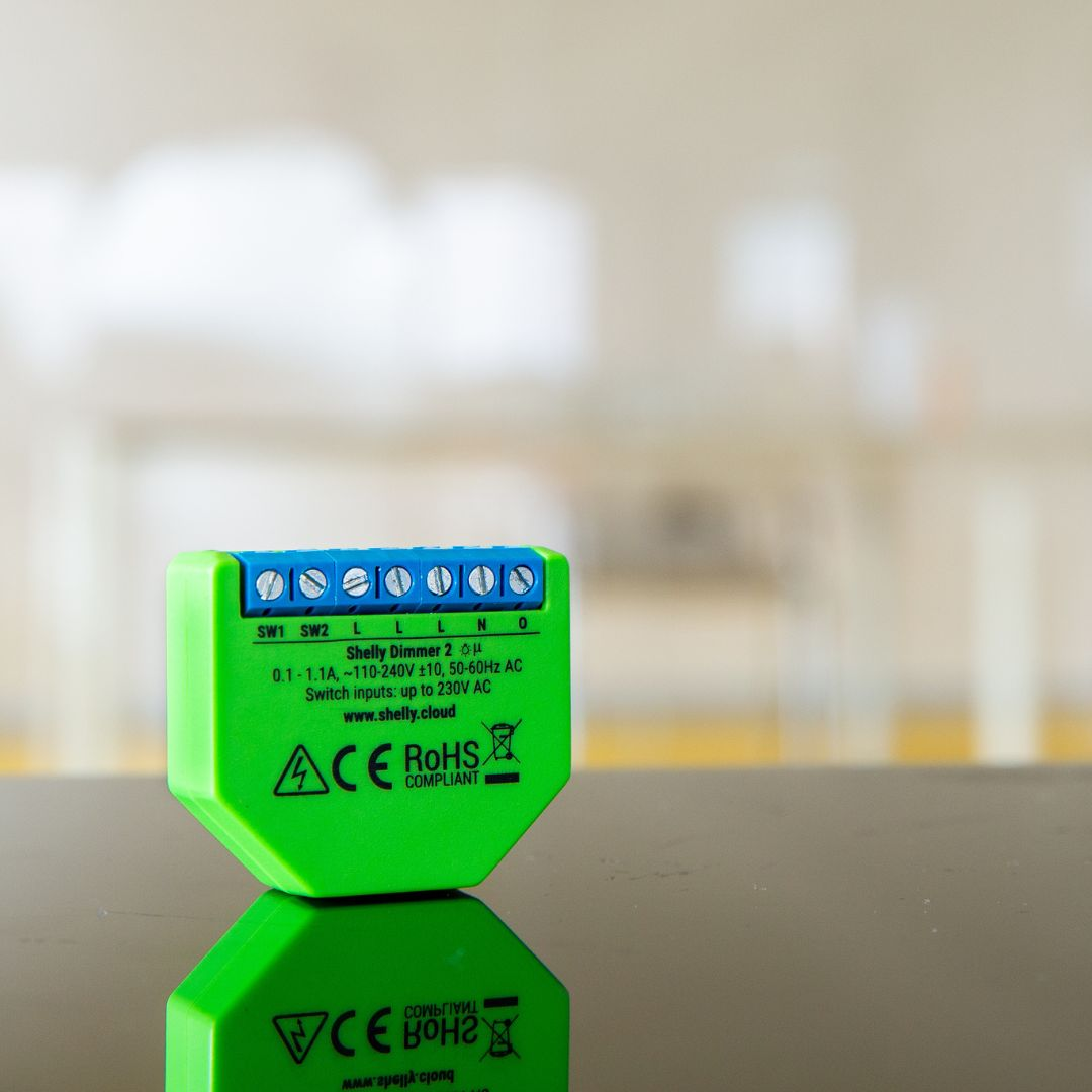 Dispositivo verde Shelly Dimmer 2 con terminali blu su un tavolo riflettente.