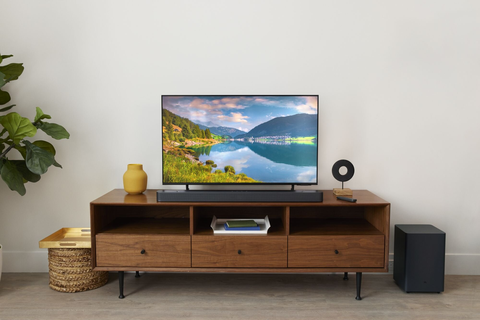 Drewniany stolik pod telewizor z telewizorem wyświetlającym krajobraz. Po lewej kosz ze złotą tacą.