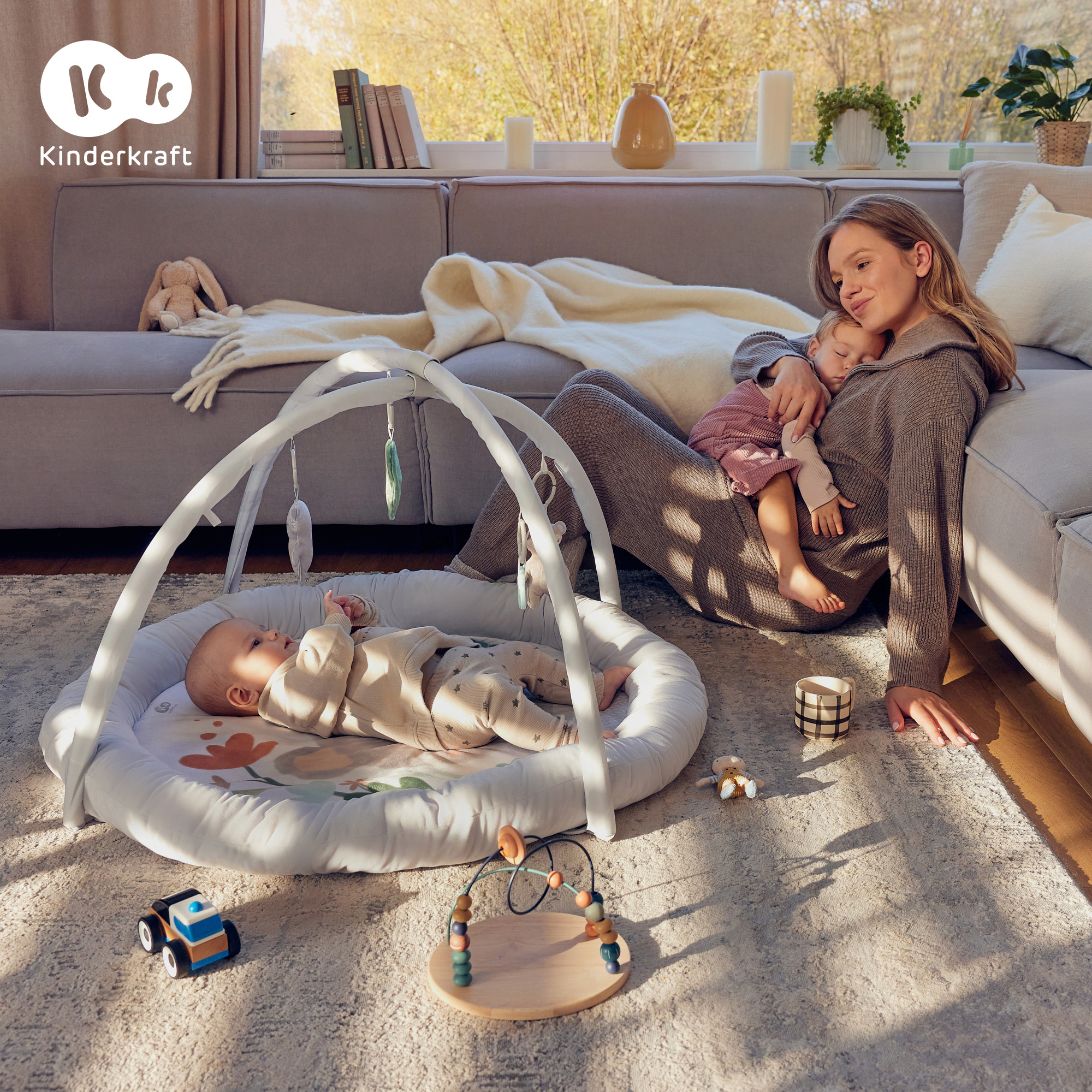 Eine Frau kuschelt mit ihrem schlafenden Kind auf dem Sofa, während ein Baby in einer Spielmatte liegt.