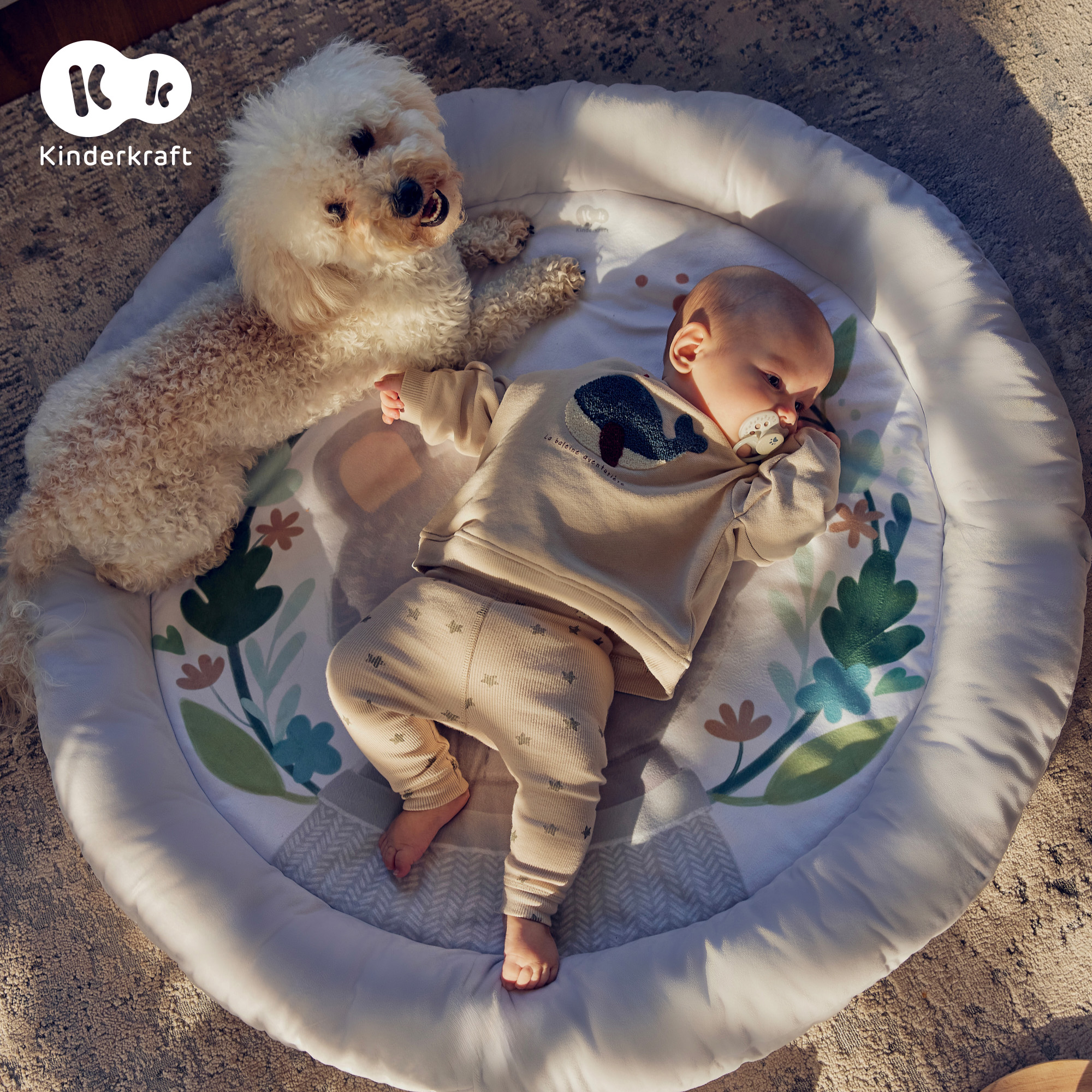 Ein weißer Hund liegt neben einem Baby auf einer Spieldecke mit Blumenmuster.