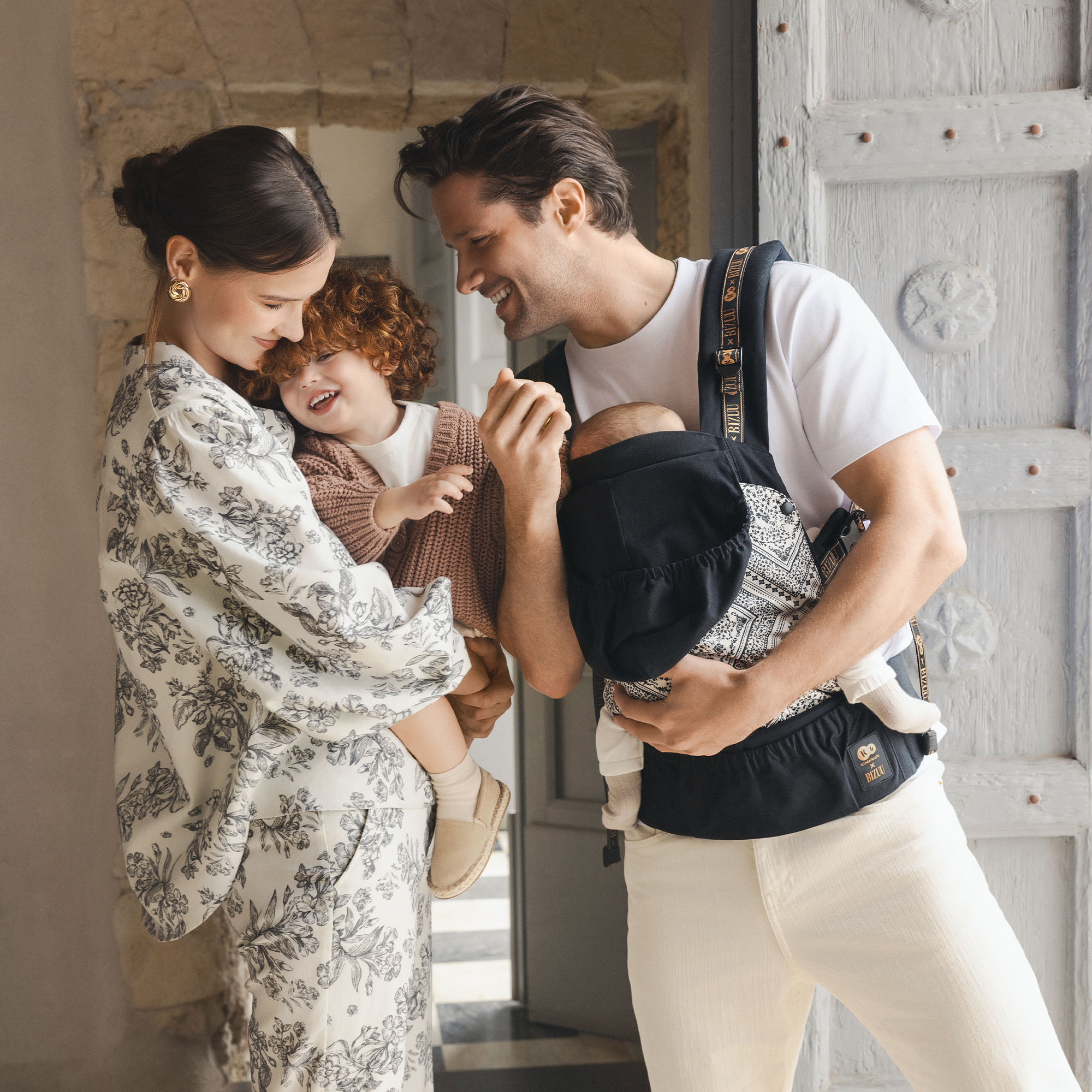 Familie mit zwei Kindern vor einer Tür. Vater trägt Baby im Tragegestell, Mutter hält Kind.