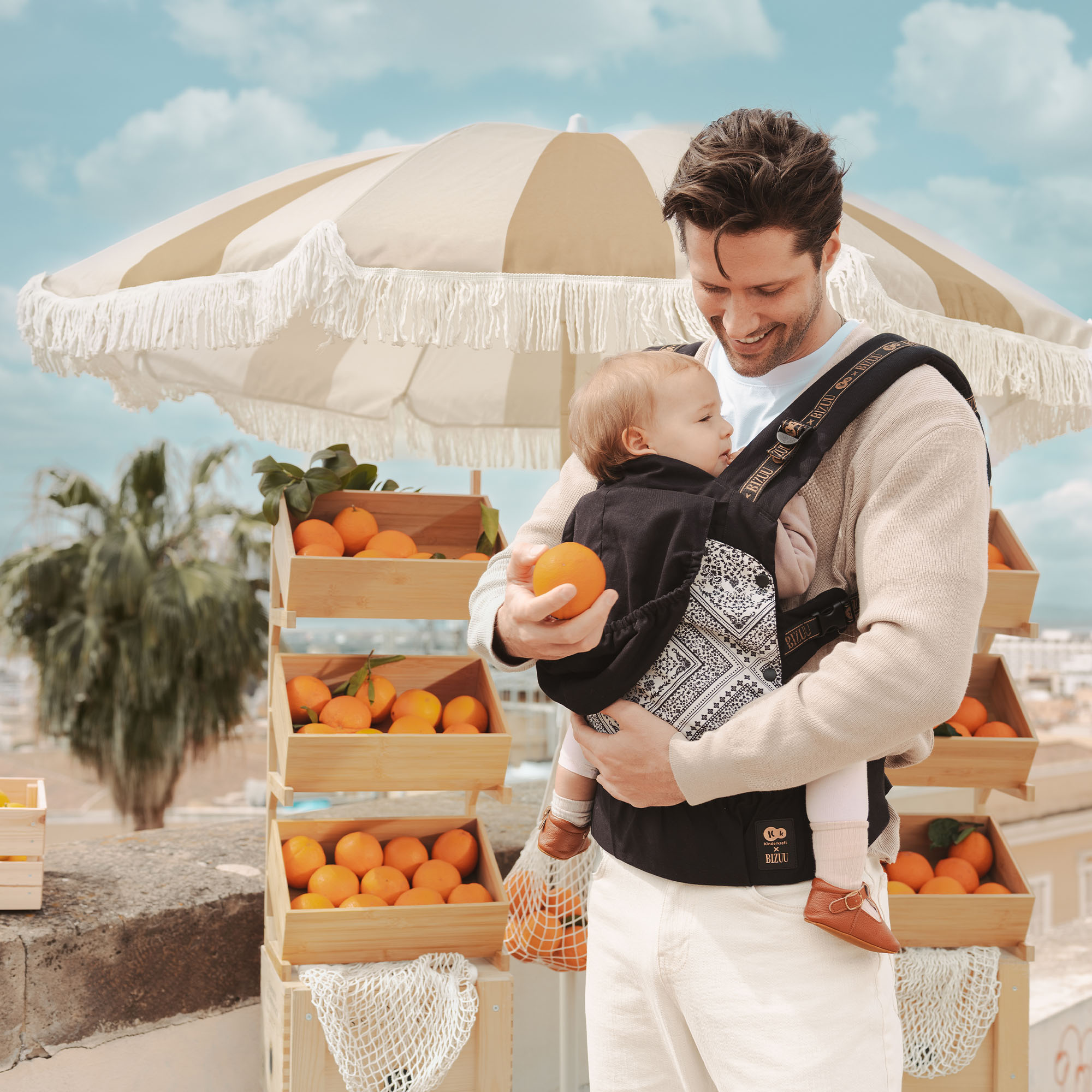 Ein Mann mit Babytrage hält eine Orange vor einem Stand mit Orangen.