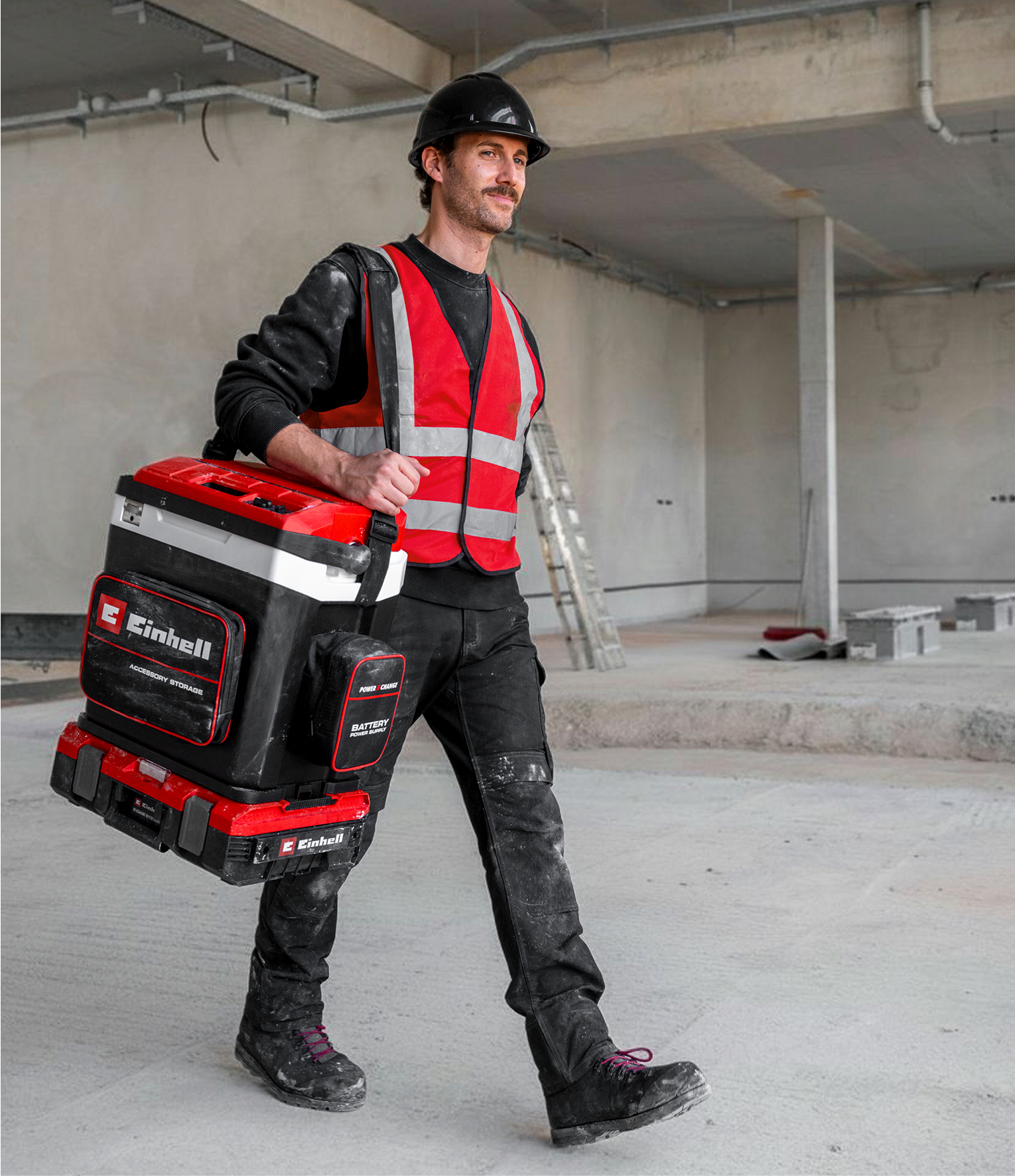 Ein Bauarbeiter mit Helm und Weste trägt eine rote und schwarze Werkzeugkiste.