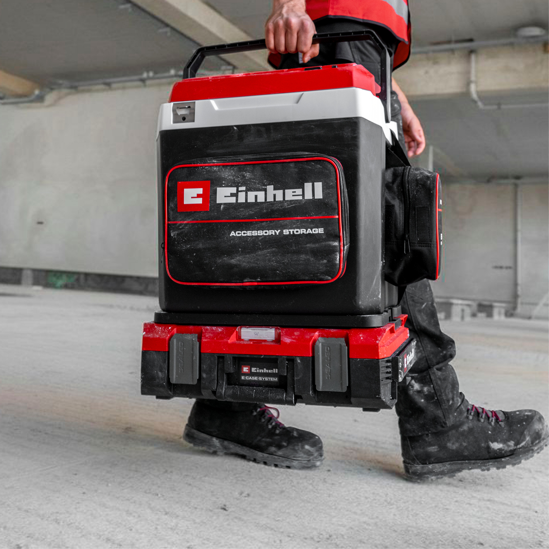 Mann trägt schwarze und rote Werkzeugkiste mit Einhell-Logo und Zubehörtasche.