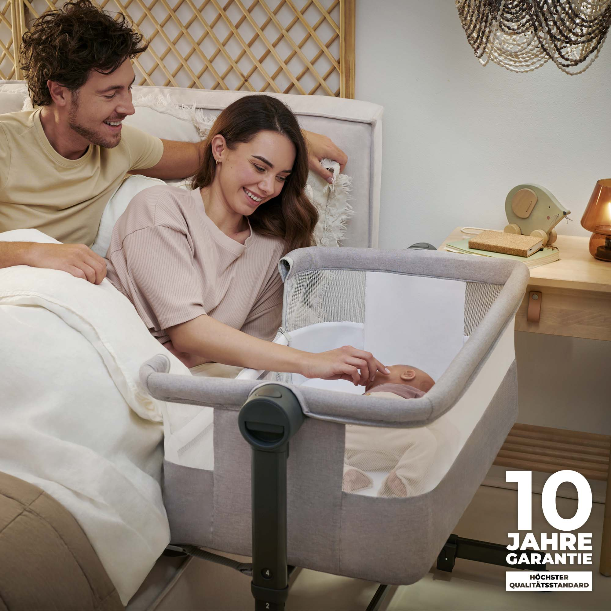 Ein Paar lächelt auf ein Baby in einem Beistellbett. 10 Jahre Garantie.