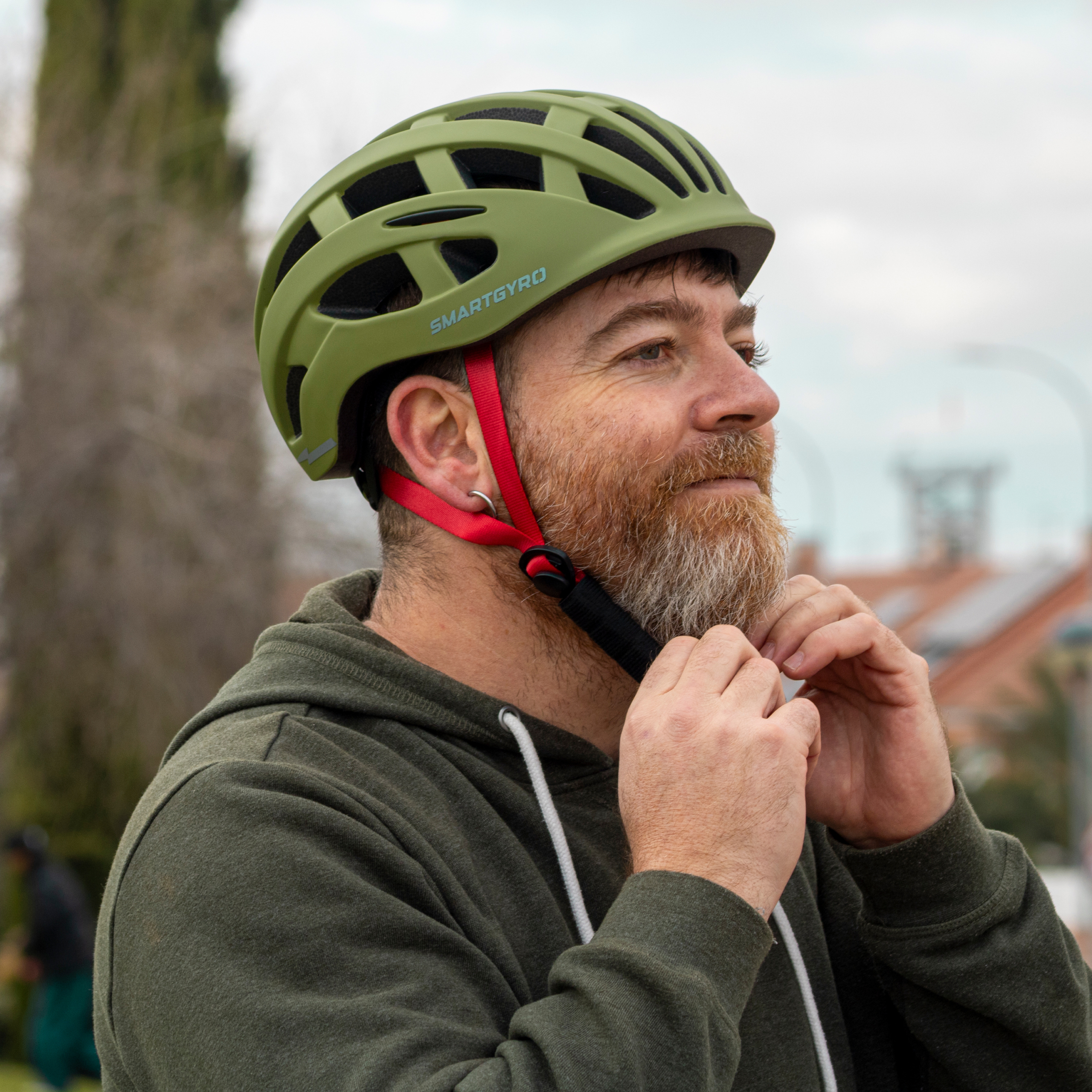 Hombre con barba abrocha casco verde con correas rojas. Fondo borroso.