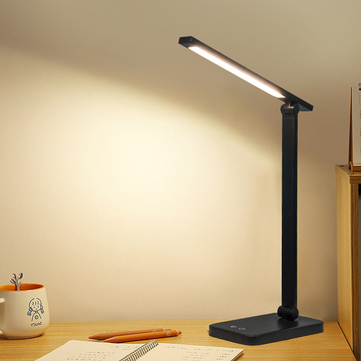Eine schwarze Schreibtischlampe leuchtet auf einem Holztisch neben einer Tasse und einem Notizbuch.