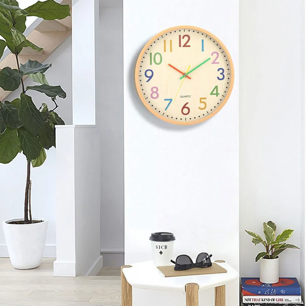 Eine Wanduhr mit bunten Zahlen hängt an einer weißen Wand neben einer Pflanze.