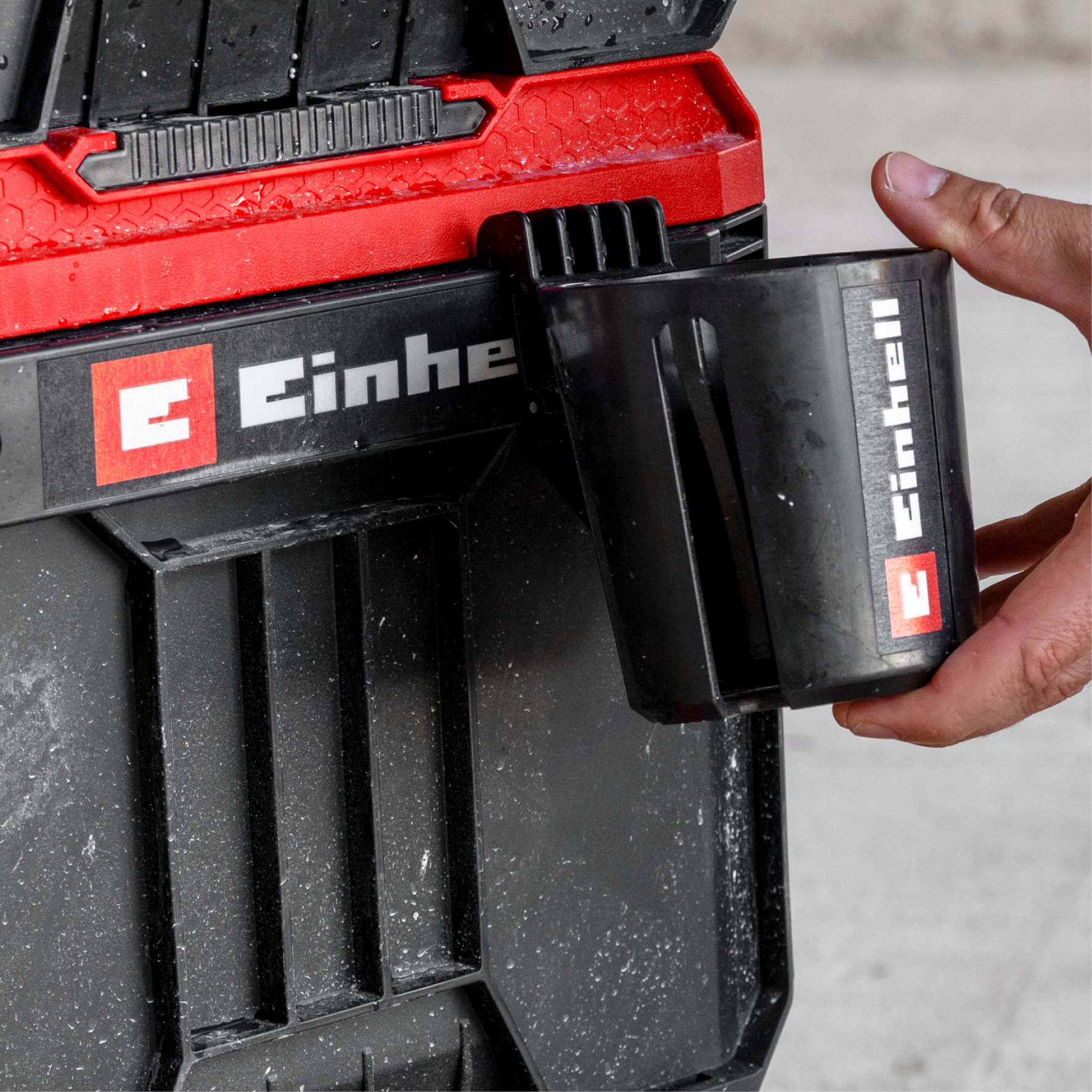 Ein Mann hält einen schwarzen Becher mit dem Einhell-Logo an eine schwarze Werkzeugkiste.