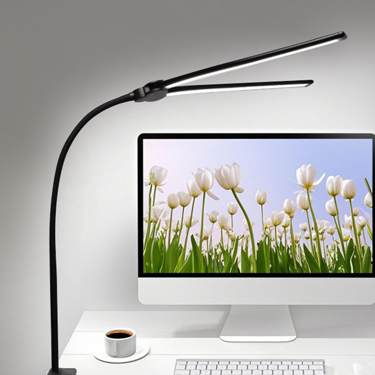 Eine schwarze Schreibtischlampe beleuchtet einen Computermonitor mit weißen Tulpen. Eine Kaffeetasse steht daneben.