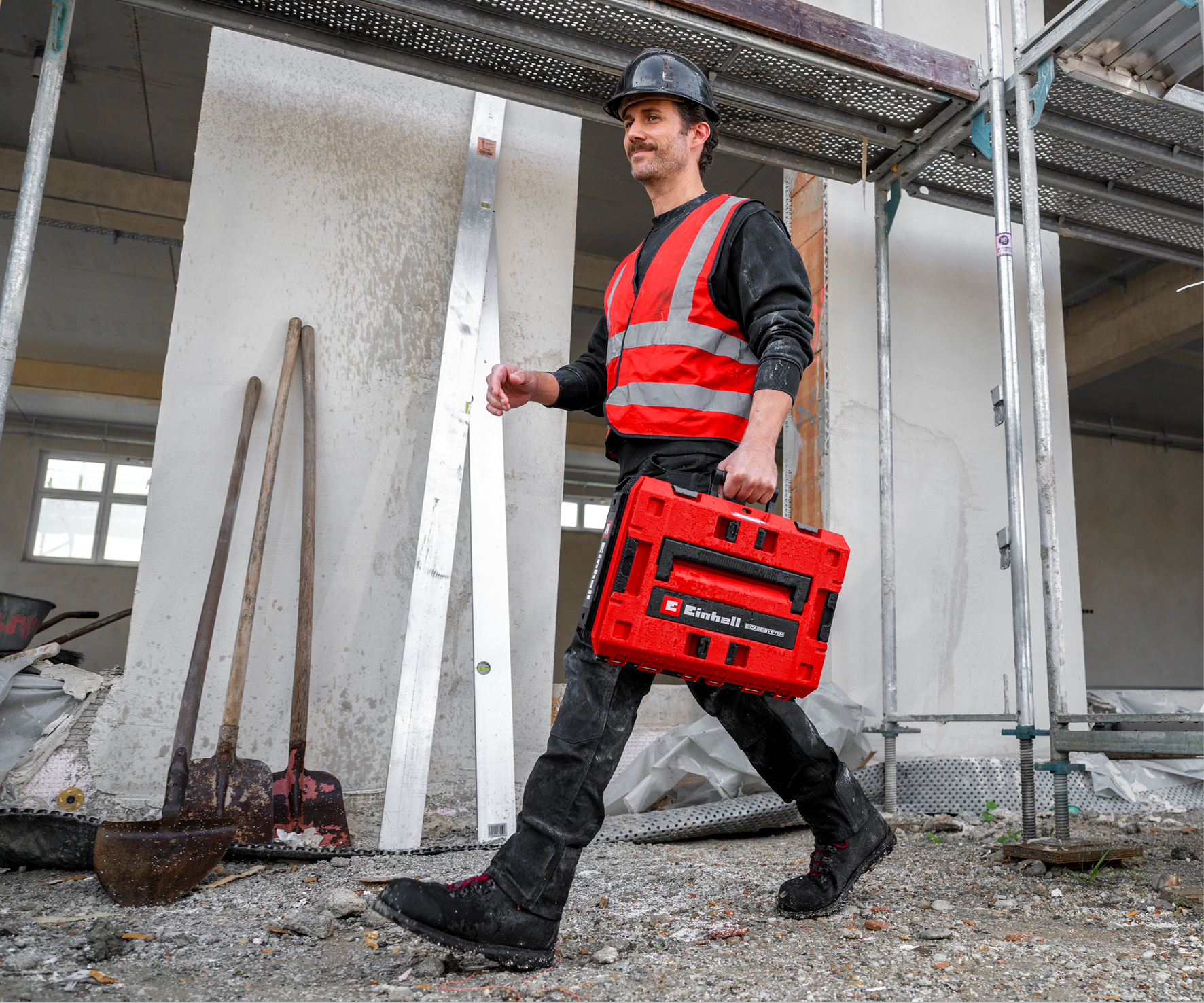 Ein Bauarbeiter in Warnweste und Helm trägt eine rote Werkzeugkiste.