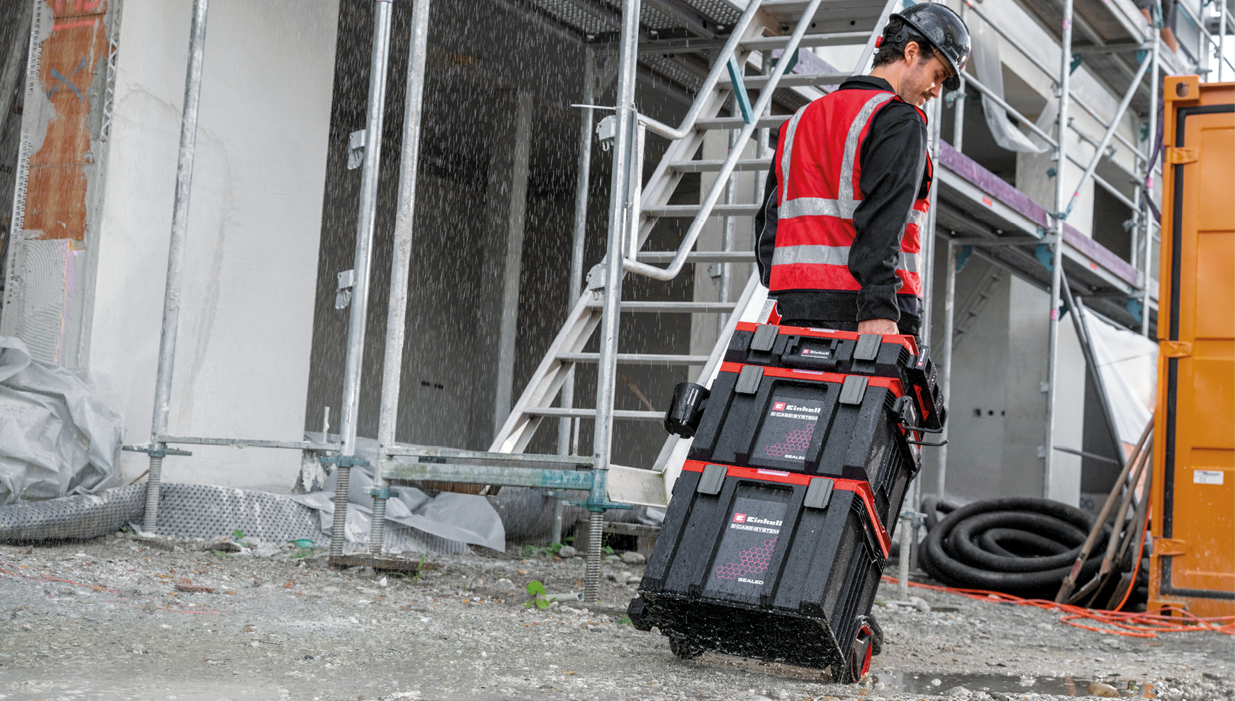 Ein Mann in Warnweste trägt Werkzeugkisten auf einer Baustelle im Regen.