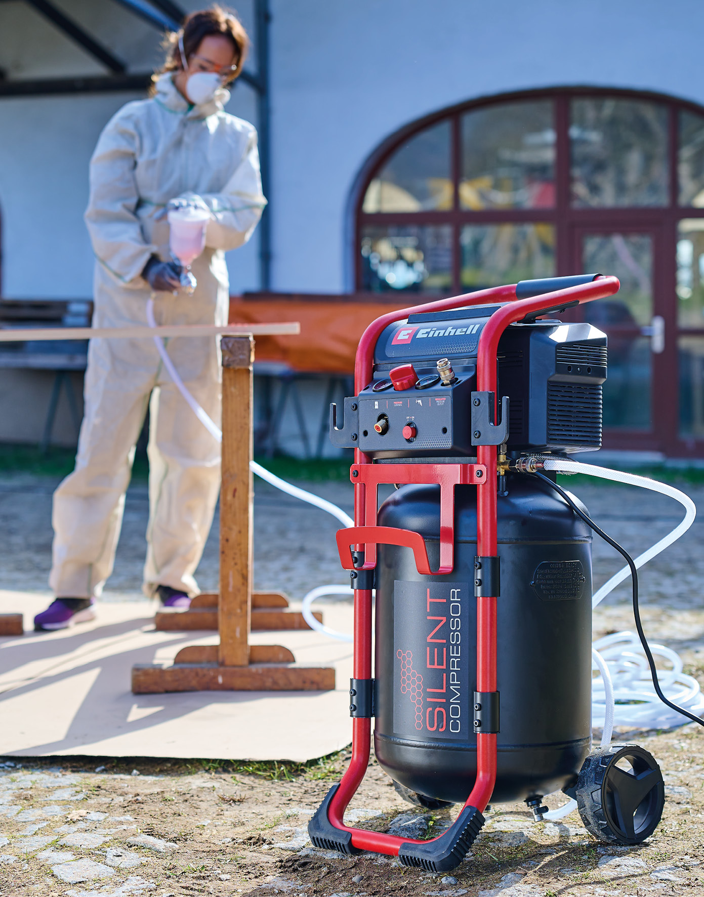 Ein Mann in Schutzkleidung sprüht mit einem Sprühgerät auf Holz. Ein Kompressor steht daneben.