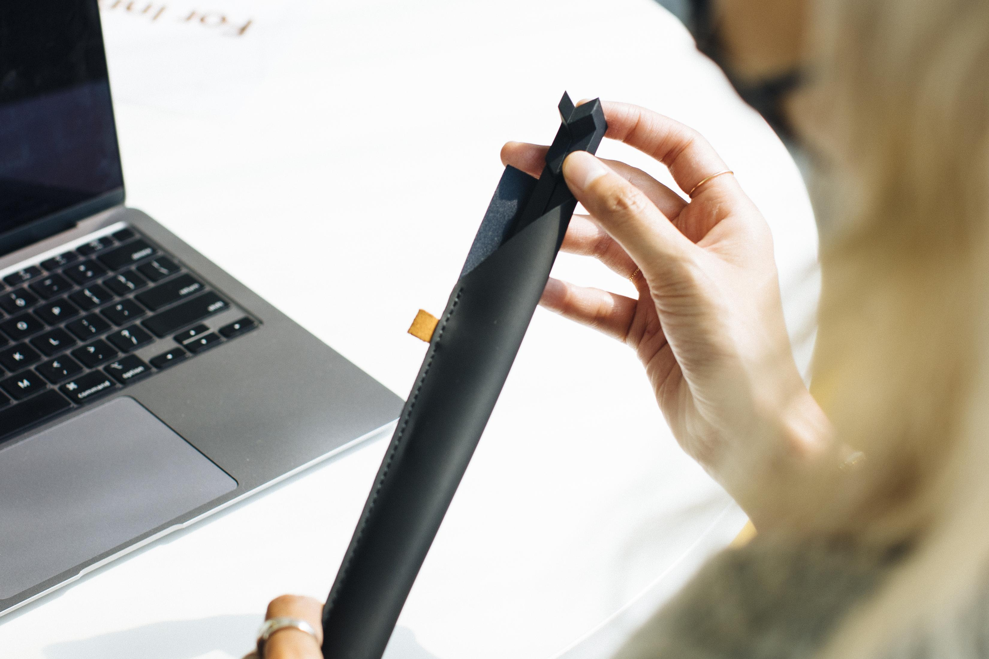 Dłoń trzyma czarne etui obok otwartego laptopa na białym stole.