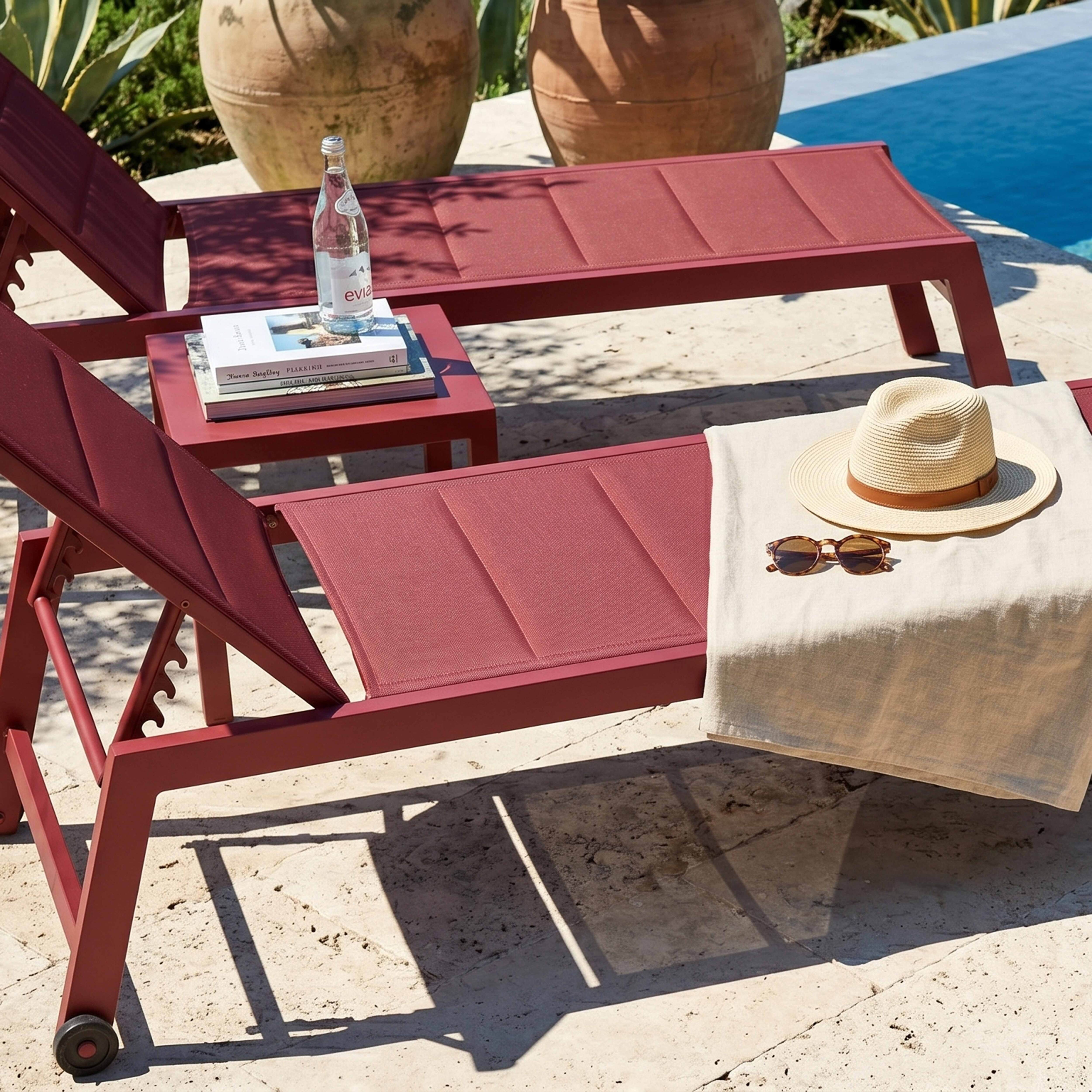 Zwei weinrote Liegestühle am Pool mit Hut, Sonnenbrille, Büchern und Wasser.