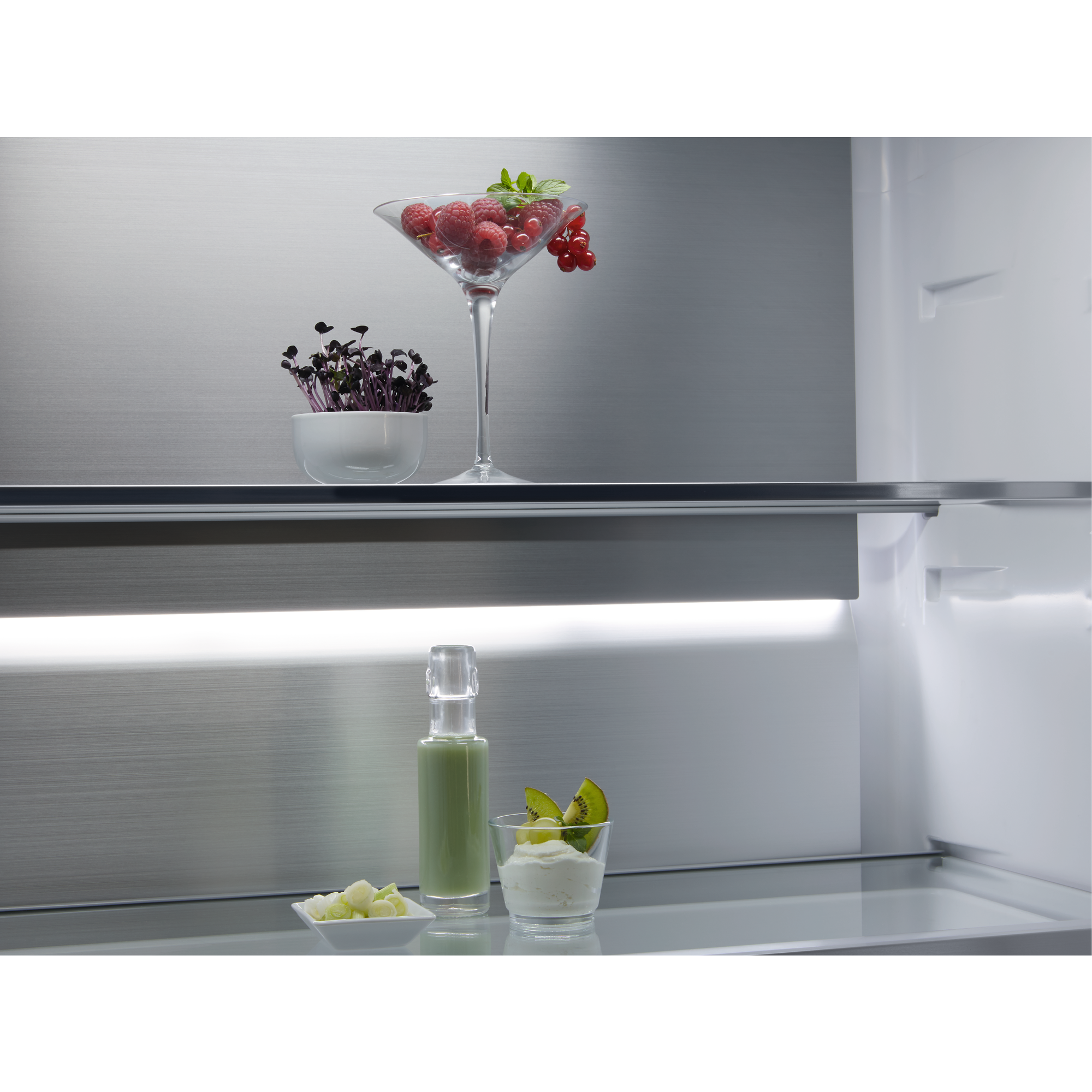 Ein Martini-Glas mit Beeren, ein Glas mit Joghurt und Kiwi, und eine Flasche grüner Flüssigkeit auf einem Regal.