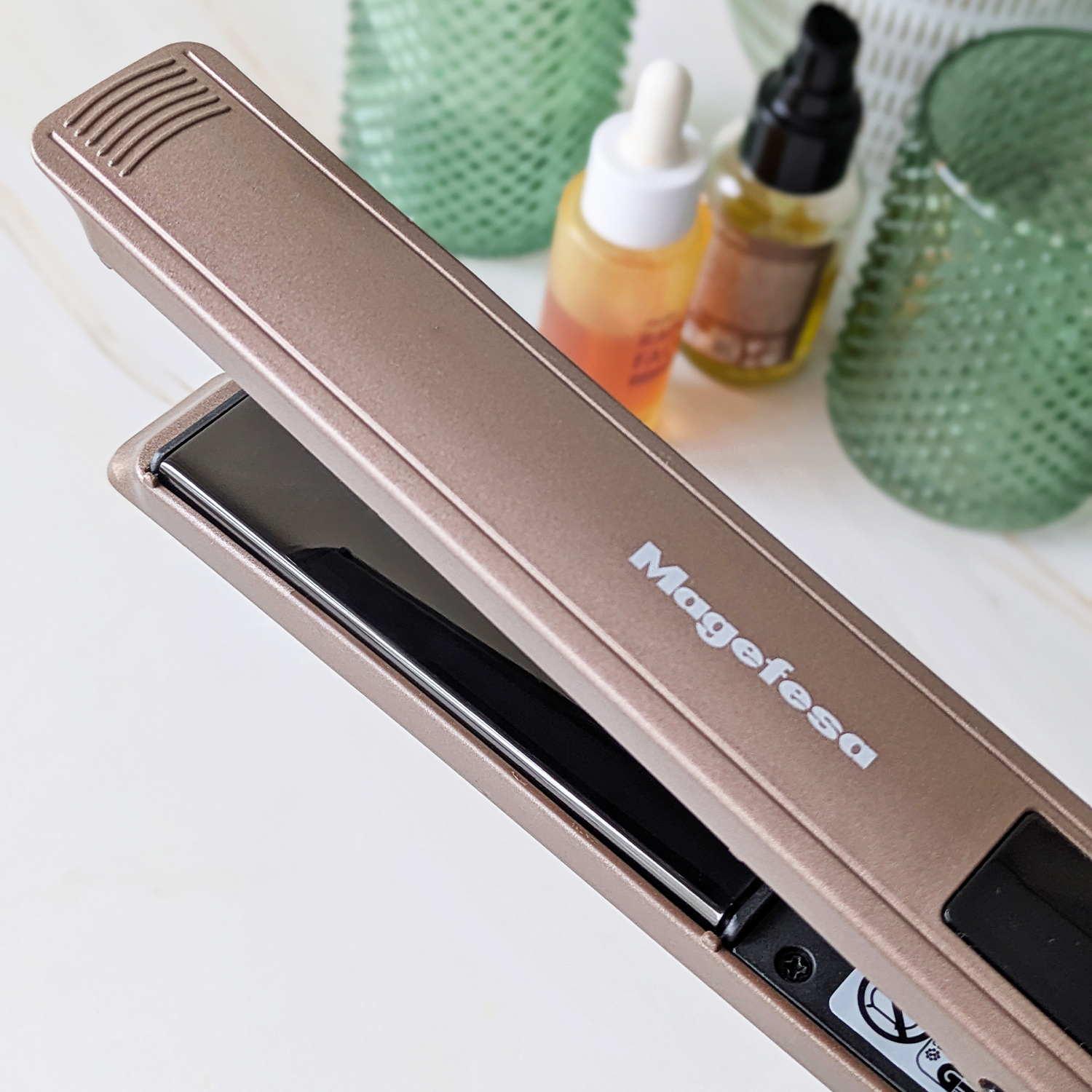 Una plancha de pelo Magefesa color oro rosa está abierta, con botellas y jarrones verdes al fondo.