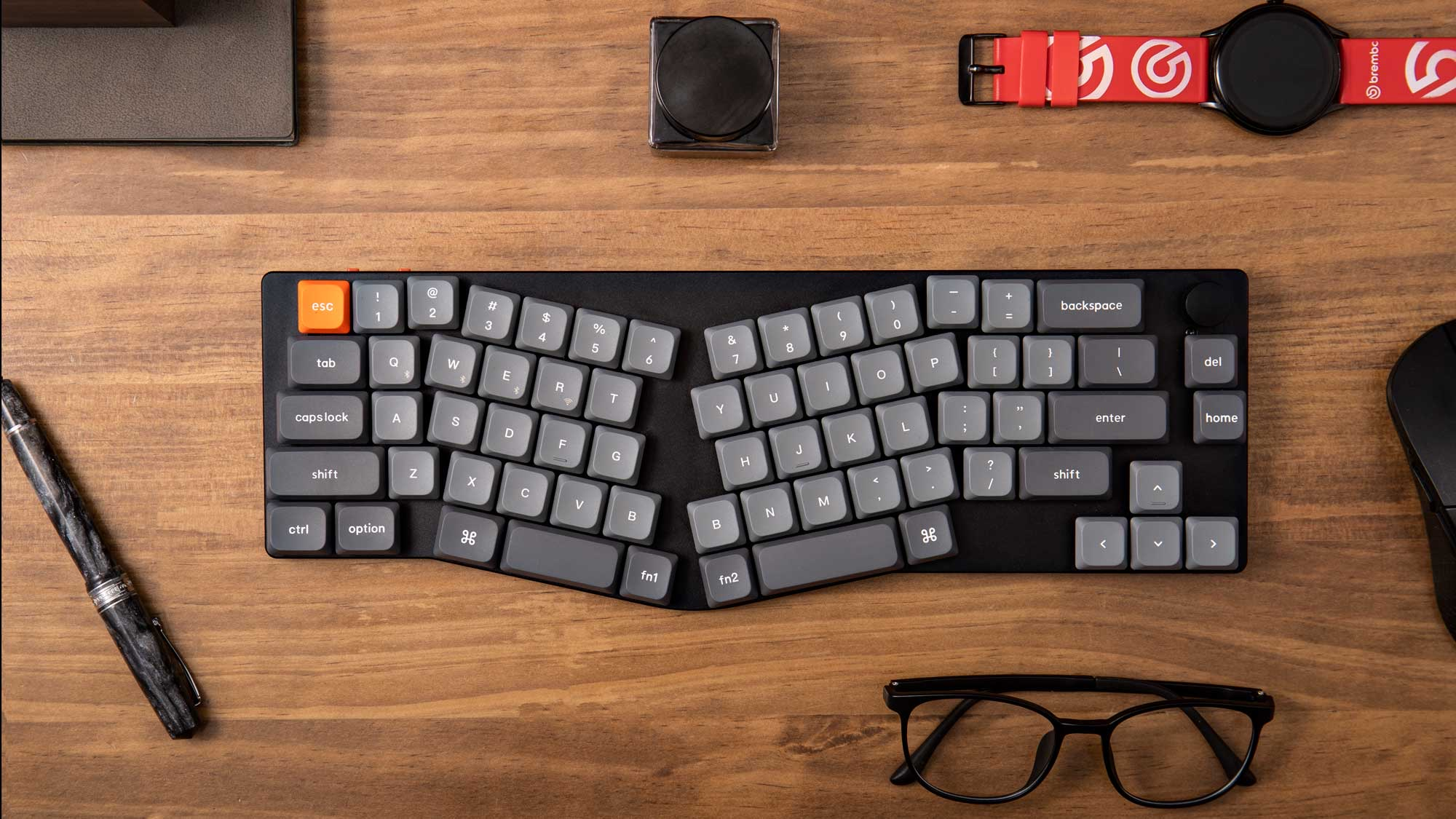 Ergonomiczna klawiatura, długopis, okulary i zegarek na drewnianym biurku.