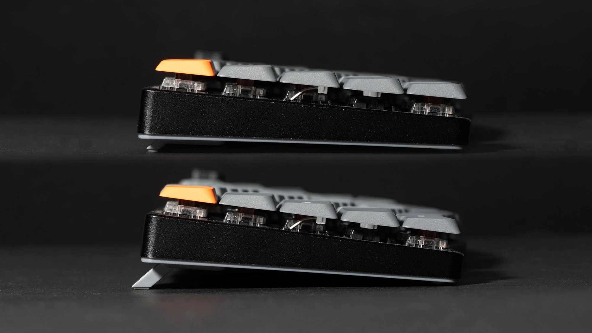 Widok z boku dwóch czarnych klawiatur z szarymi klawiszami i jednym pomarańczowym. Czarne tło.