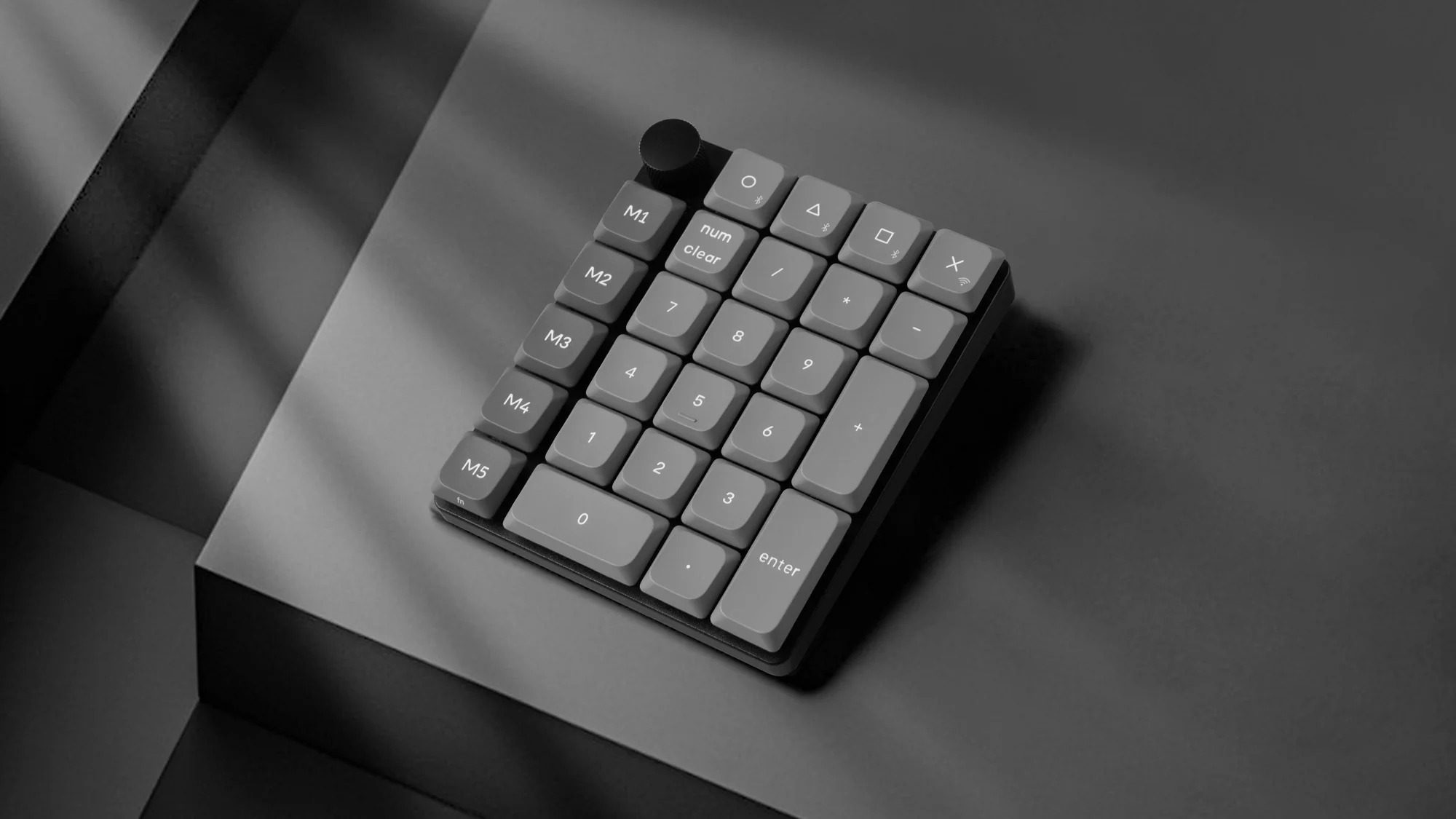 Szara klawiatura numeryczna z klawiszami M1-M5 i pokrętłem, na ciemnej powierzchni.