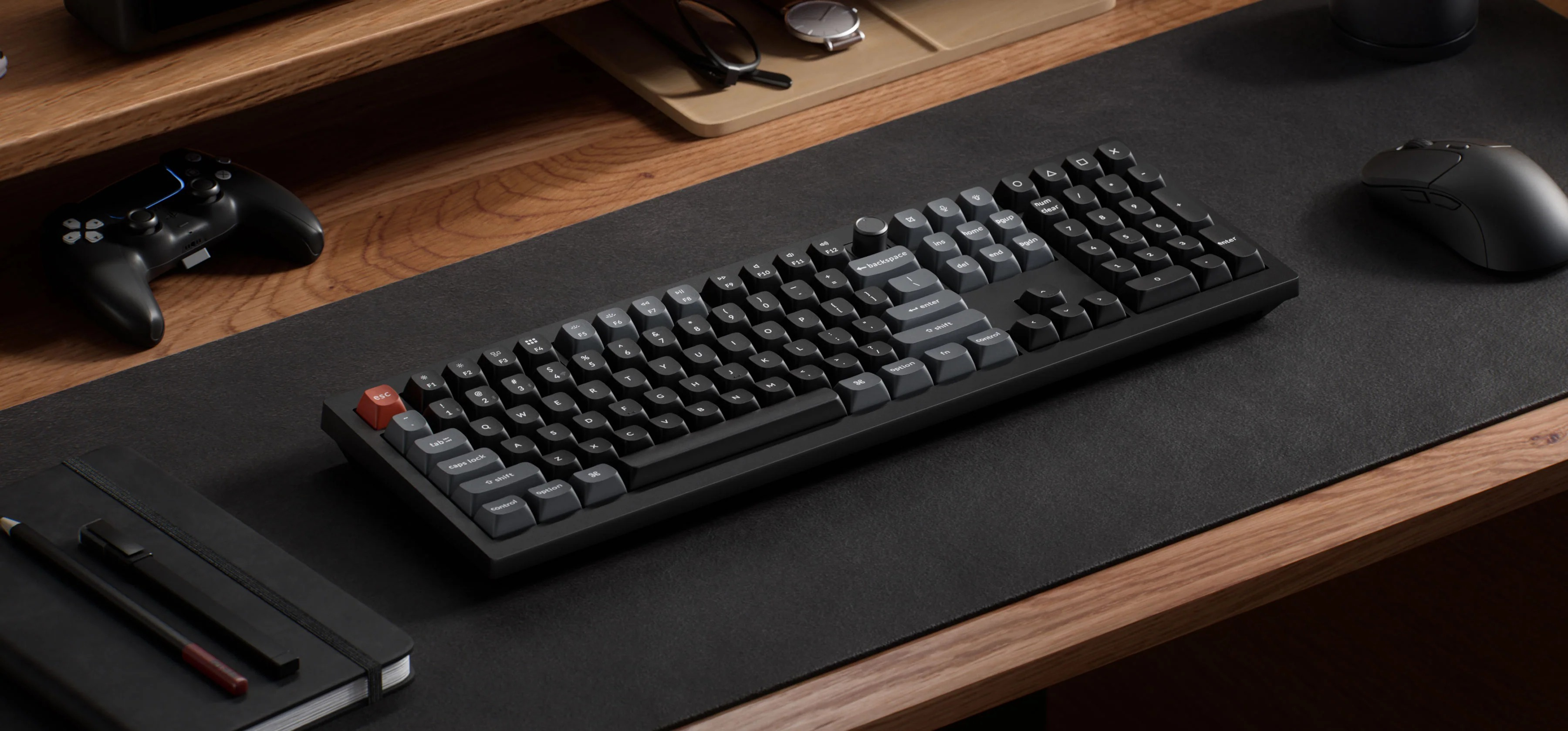 Czarna klawiatura z czerwoną klawiszem ESC, myszą, kontrolerem, notatnikiem i długopisami na biurku.