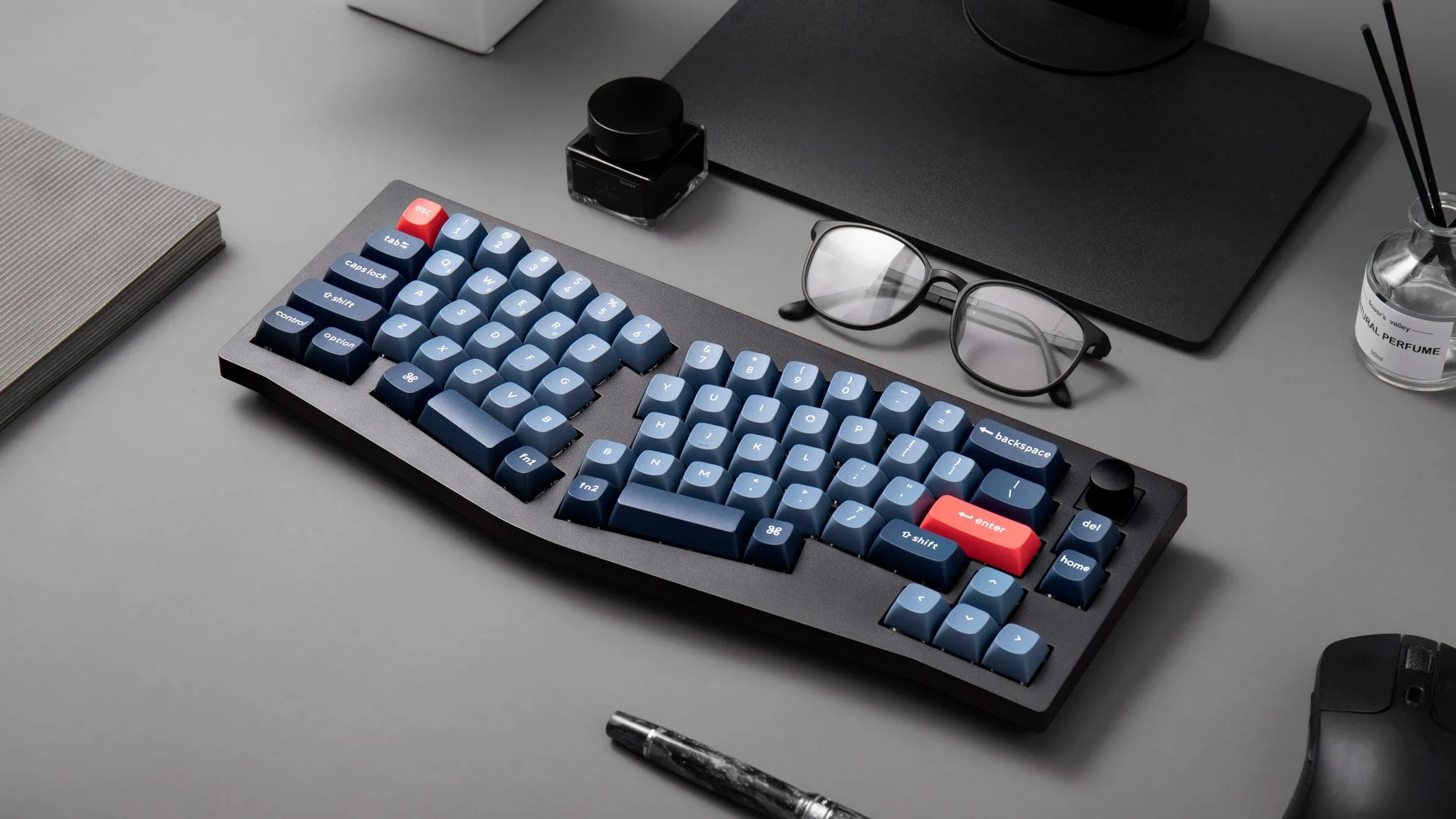 Niebieska klawiatura mechaniczna z czerwonymi akcentami na szarym biurku. Zawiera okulary, atrament i mysz.
