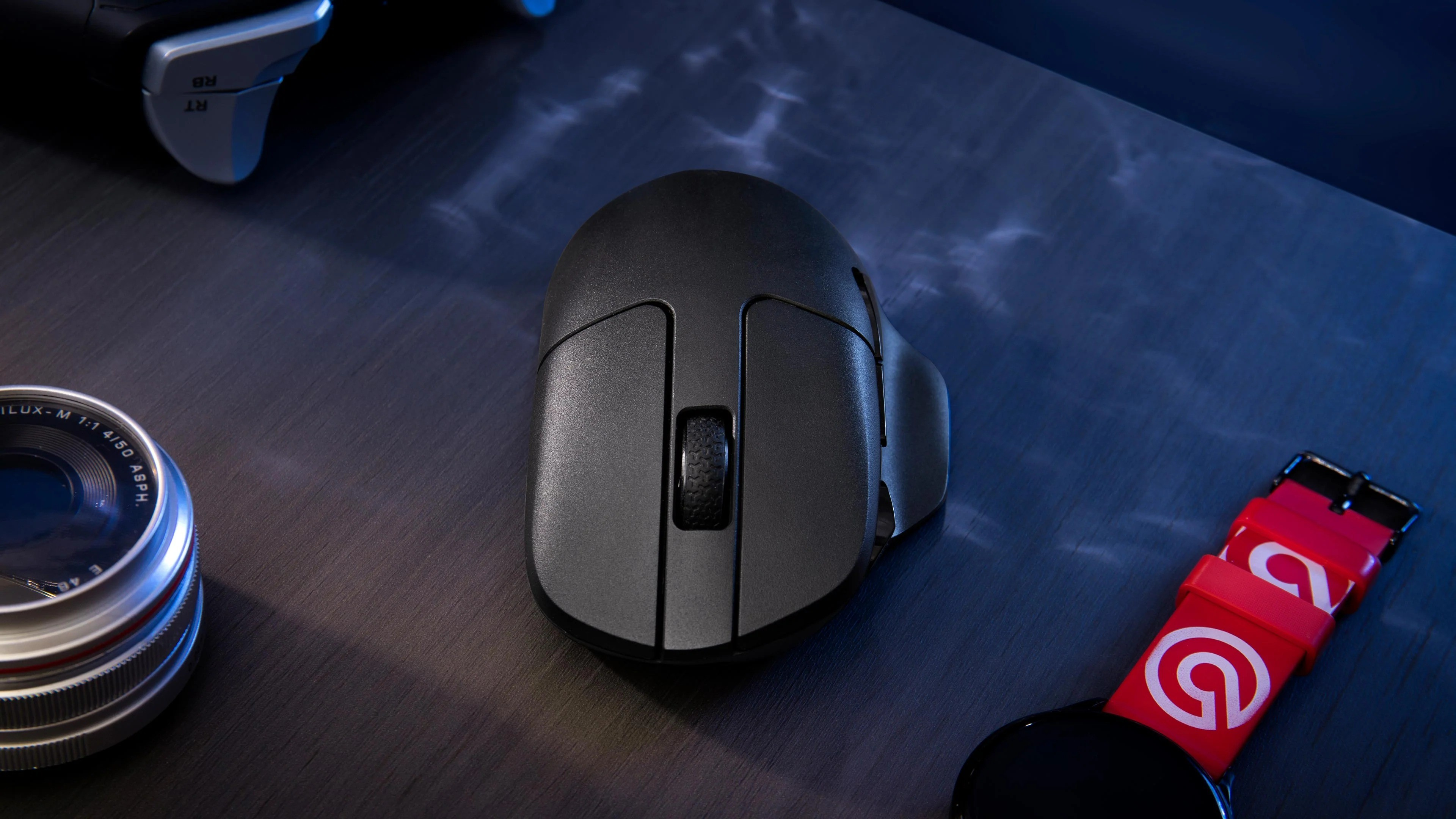 Czarna mysz gamingowa, obiektyw aparatu i czerwona opaska smartwatch na ciemnym drewnie.