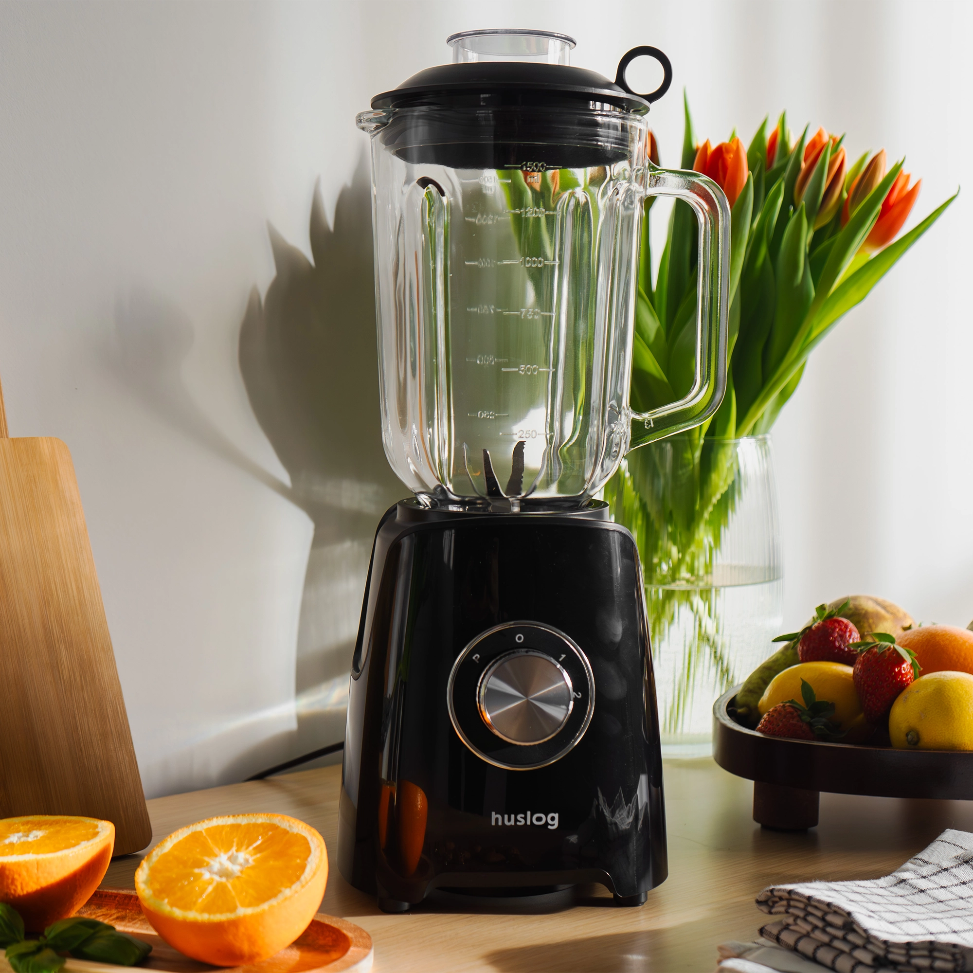 Czarny blender ze szklanym dzbankiem, obok pomarańczy, jagód i tulipanów.