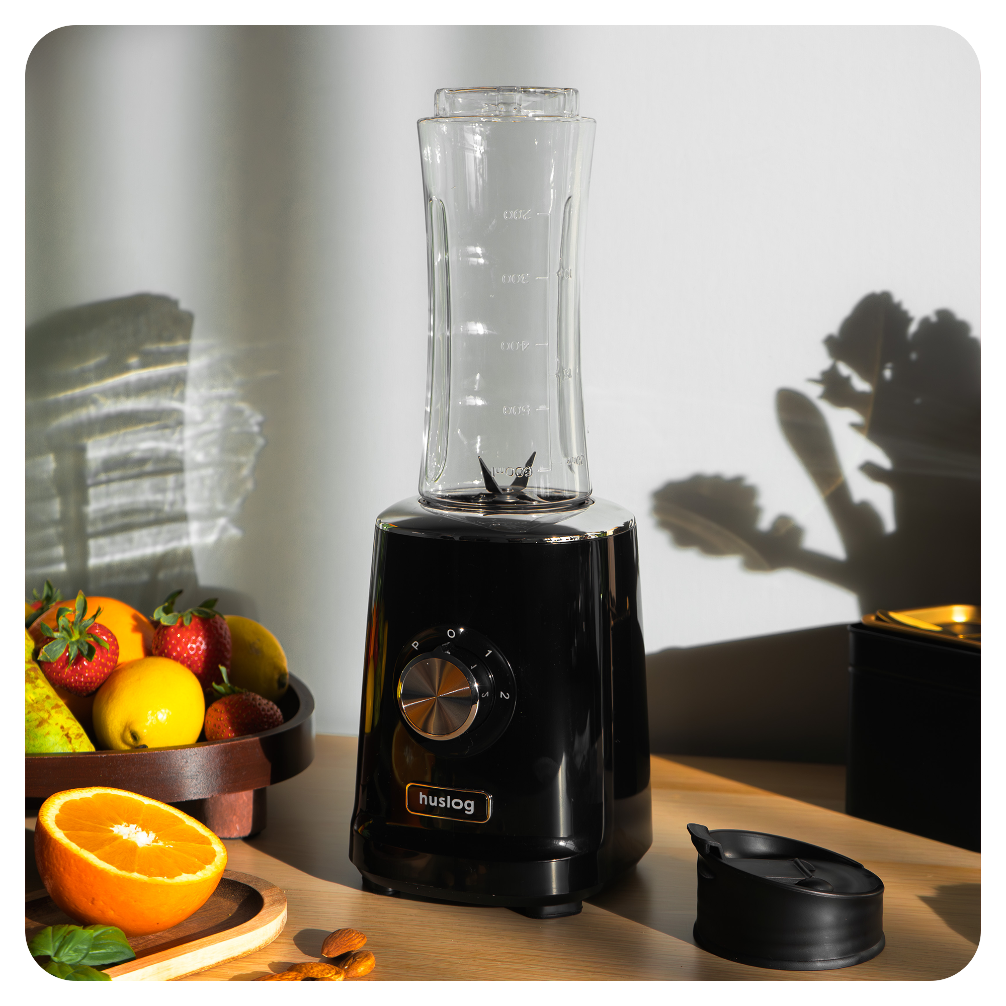 Czarny blender z szklanym naczyniem, obok miska z owocami, truskawkami i cytrynami.