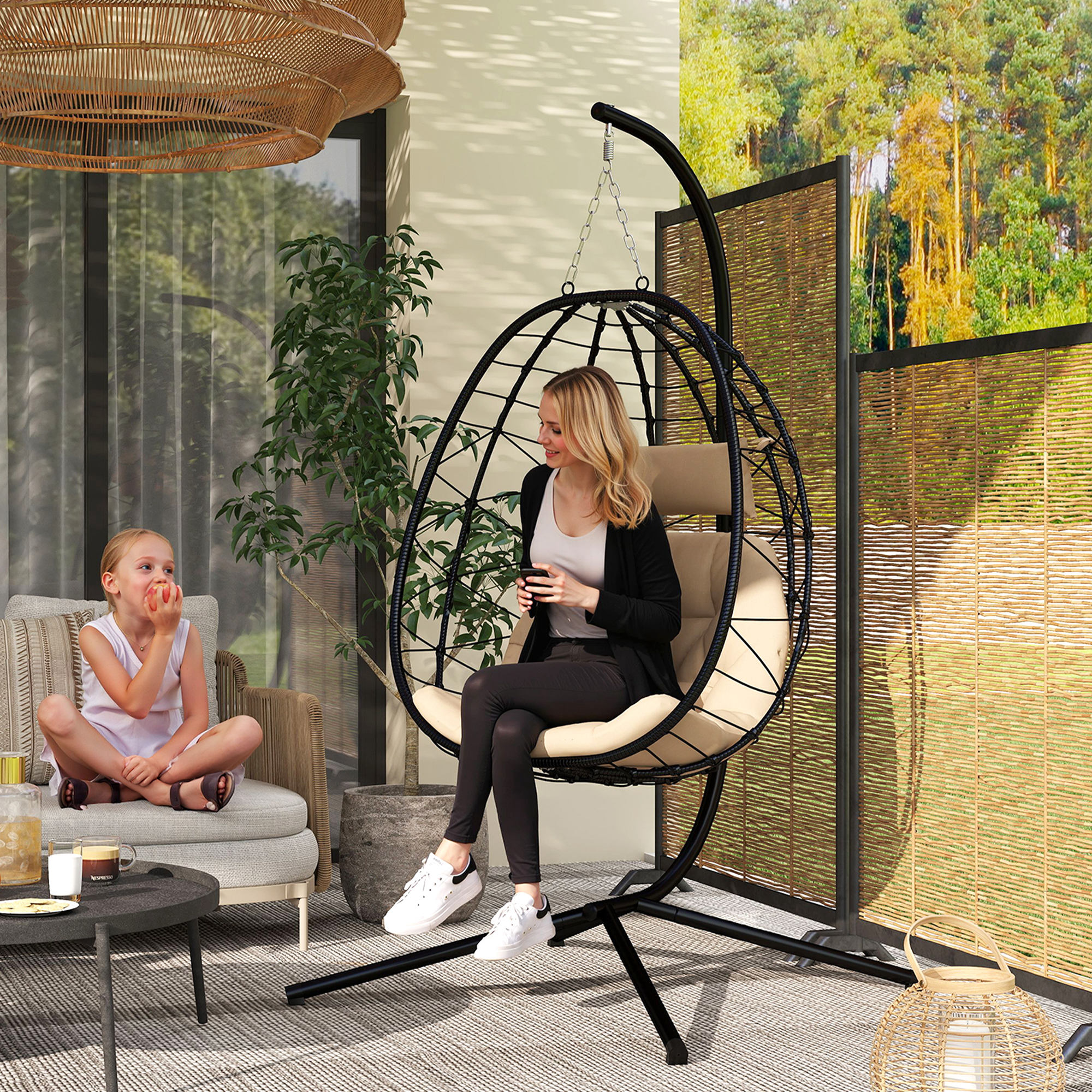 Mujer en silla colgante, niña en sofá, patio con plantas y fondo de árboles.