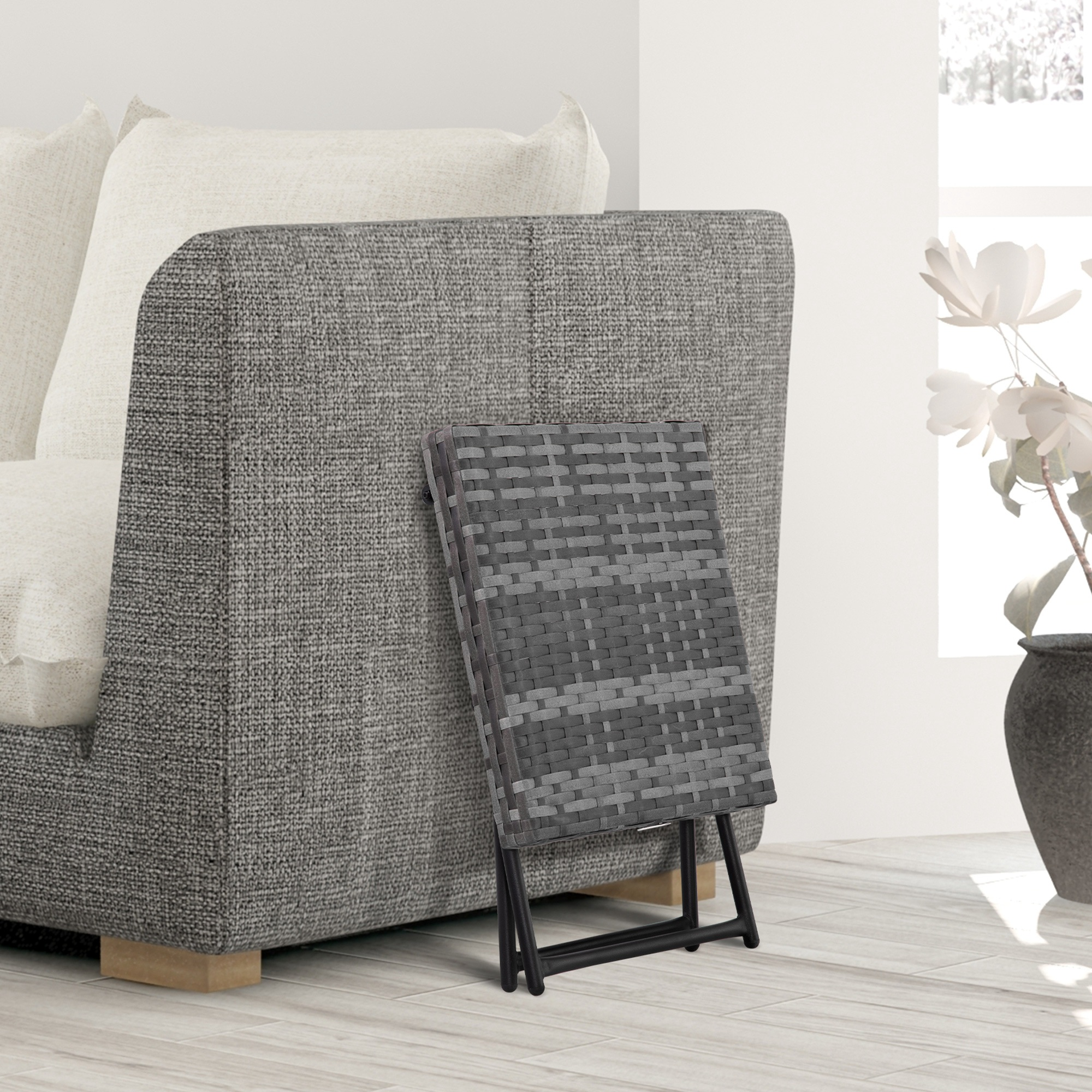 Tavolo pieghevole in rattan grigio appoggiato su un divano grigio con cuscini bianchi.