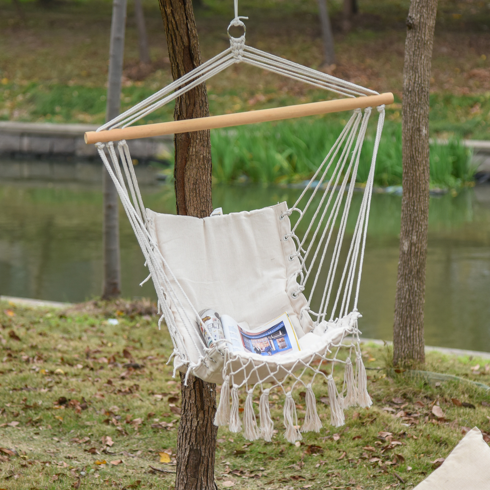 Una silla hamaca de macramé blanca con borlas cuelga de un árbol junto a un estanque.