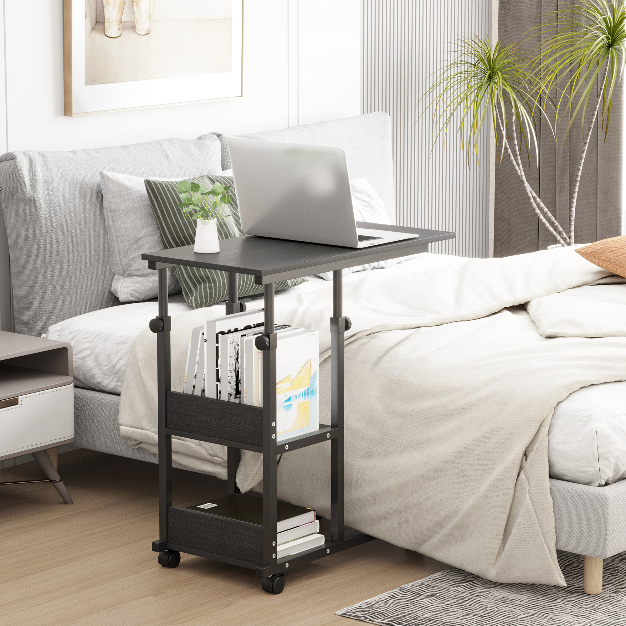 Una mesa portátil con ruedas junto a una cama. Libros y una planta sobre ella.