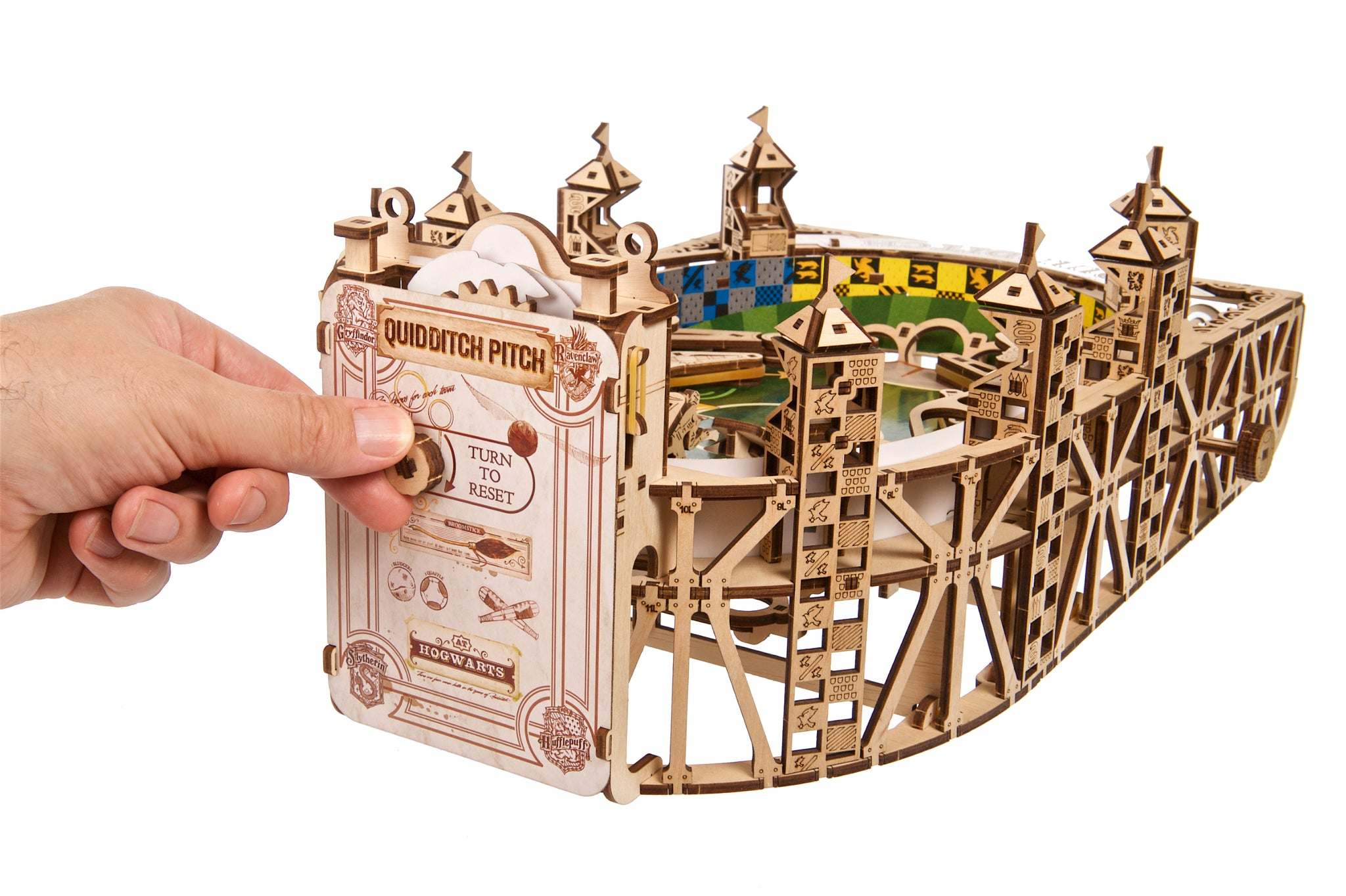 Eine Hand dreht einen Holzknebel an einem Quidditch-Spielfeldmodell mit Hogwarts-Wappen.