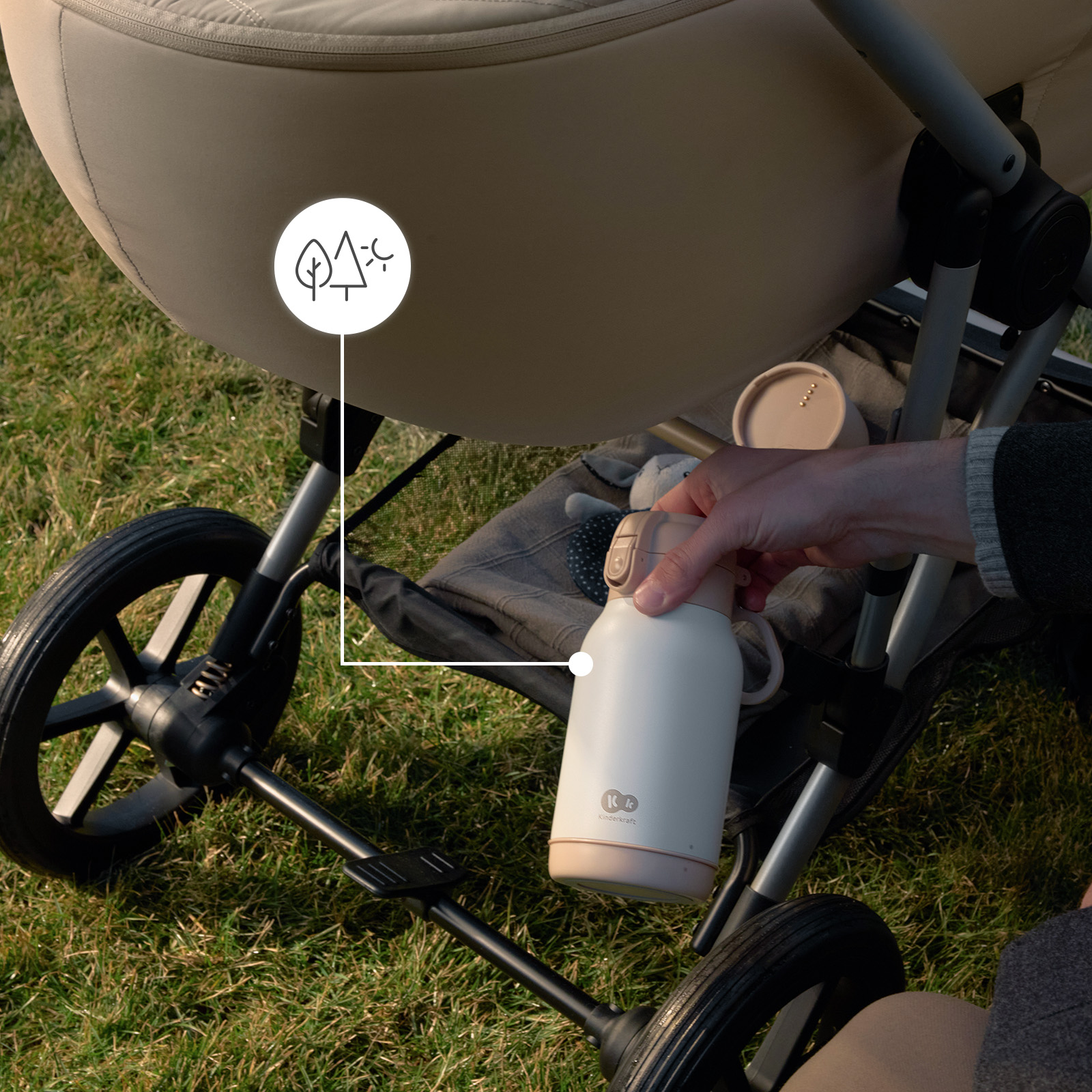Eine Hand greift nach einer weißen Thermoskanne neben einem Kinderwagen im Gras.
