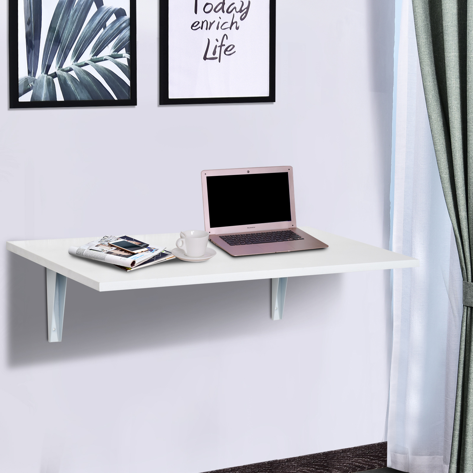 Un escritorio blanco de pared con laptop, taza y revistas. Cerca hay una ventana con cortinas.