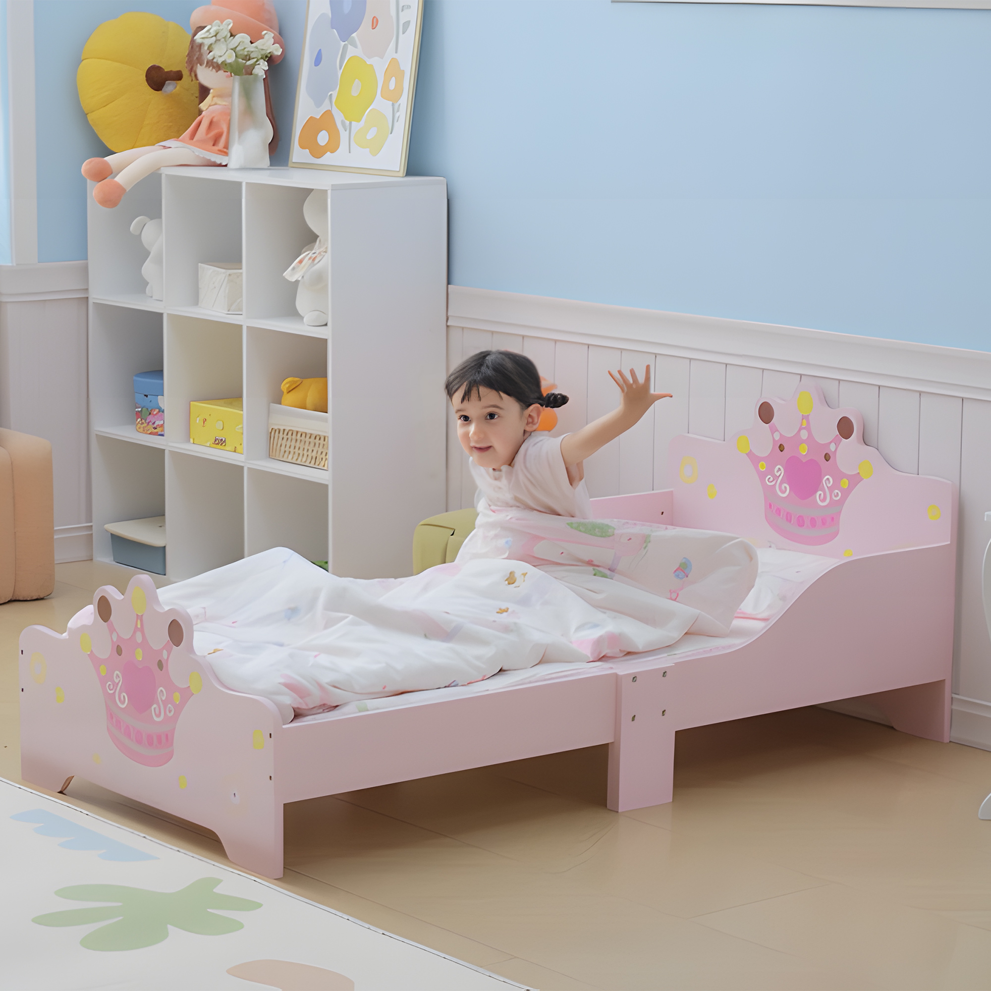 Una niña pequeña se sienta en una cama rosa con temática de corona y ropa de cama blanca.